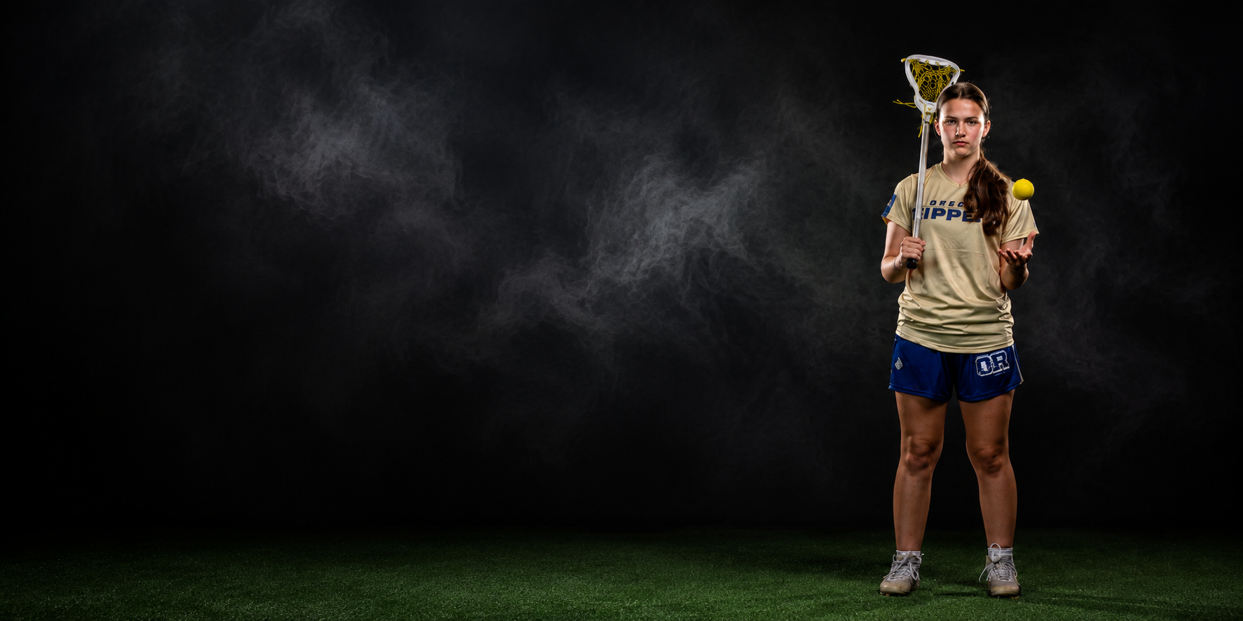 A young woman in sports gear practicing lacrosse, holding a lacrosse stick and a yellow ball, standing on artificial turf with a dark background.