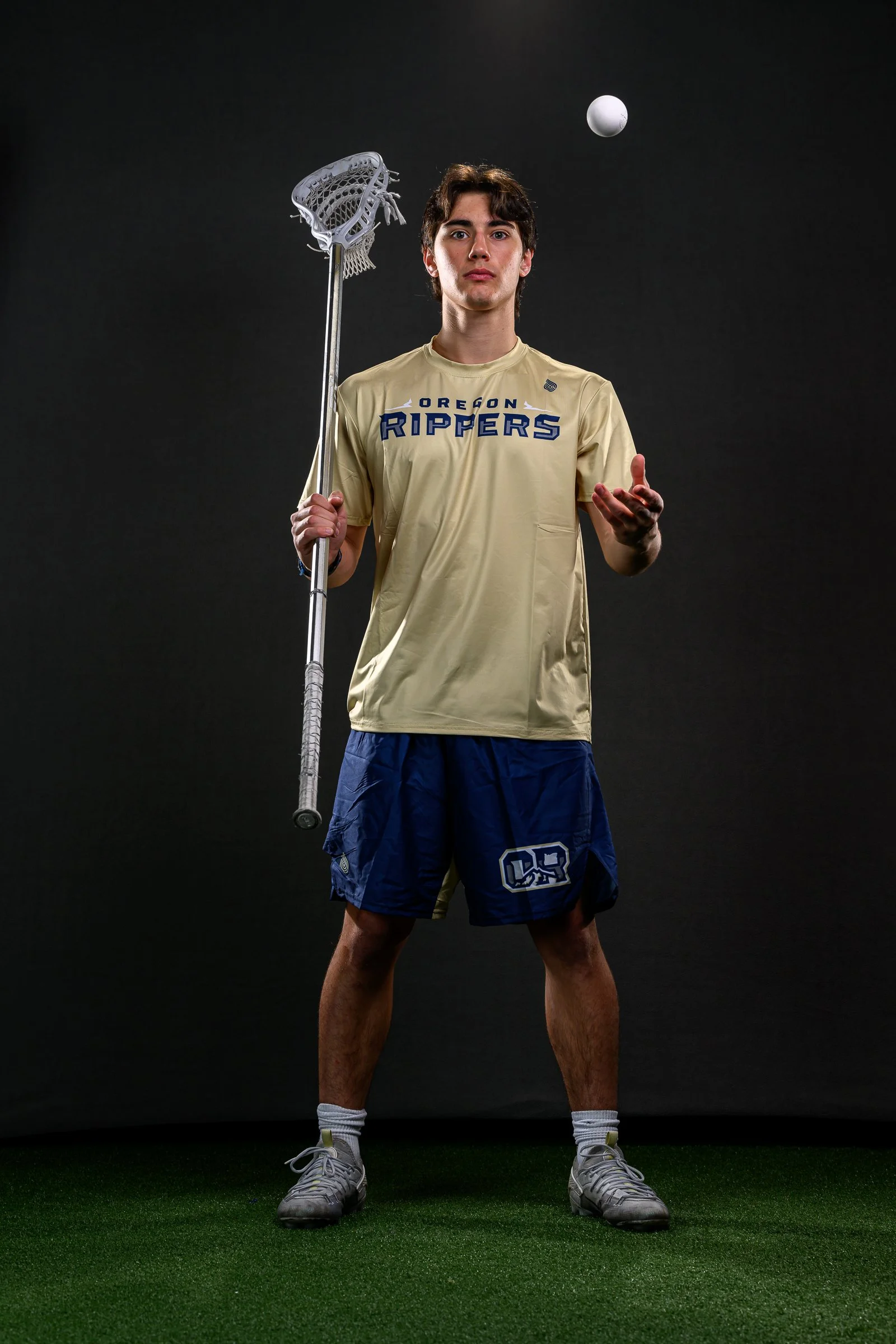 Young male lacrosse player standing on a green turf field, holding a lacrosse stick in one hand, wearing a beige Oregon Rippers jersey, navy shorts, white socks, and grey sneakers, with a lacrosse ball floating above his head against a black background.
