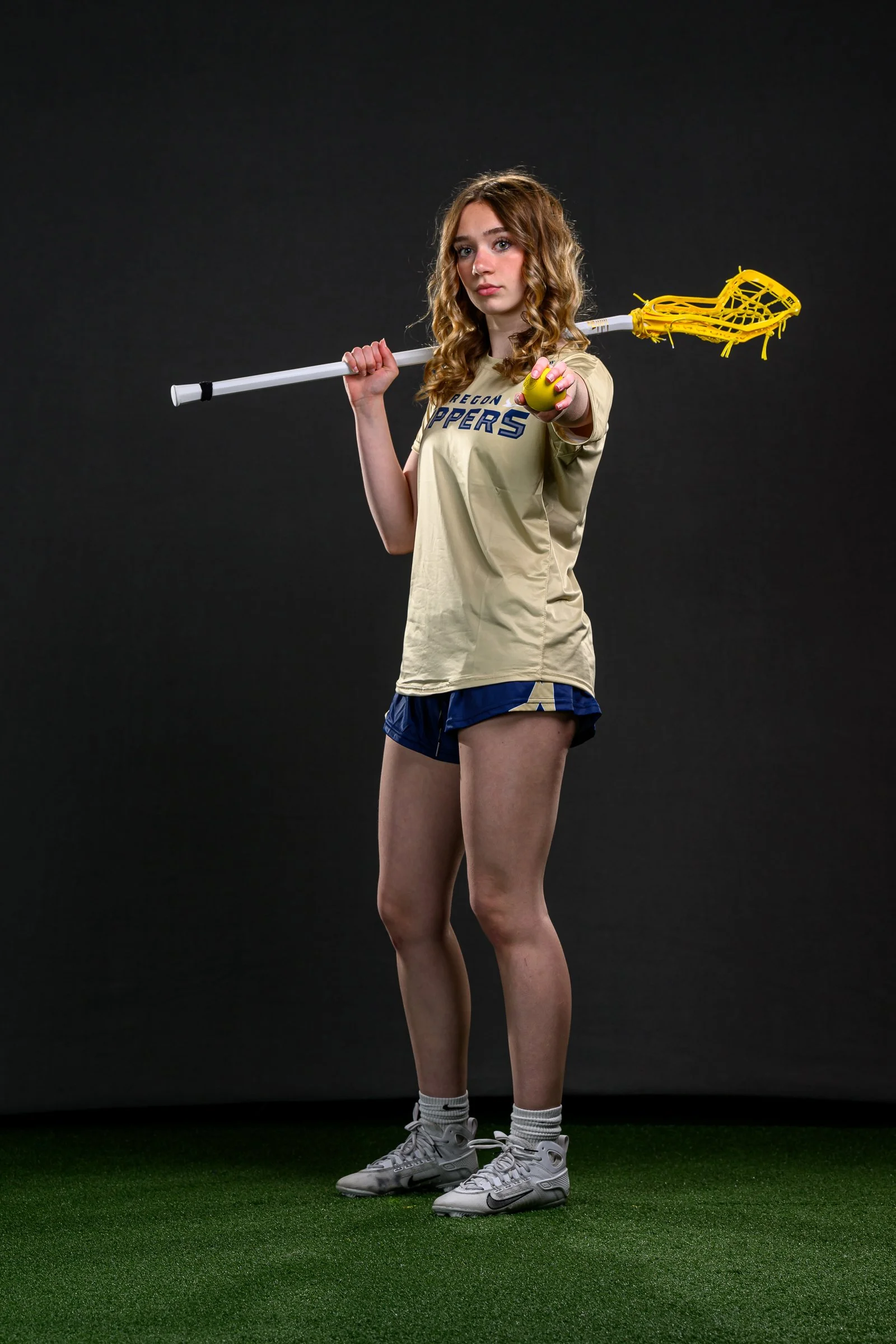 A young woman holding a lacrosse stick over her shoulder and pointing forward, wearing a sports jersey and shorts, standing on artificial turf against a black background.