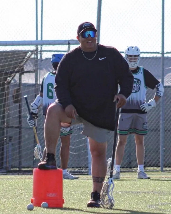 Coach Koda_Wataka Robinson on a lacrosse field standing with one foot on an orange bucket, holding a lacrosse stick and leading a training drill, with young players in uniforms standing behind him.
