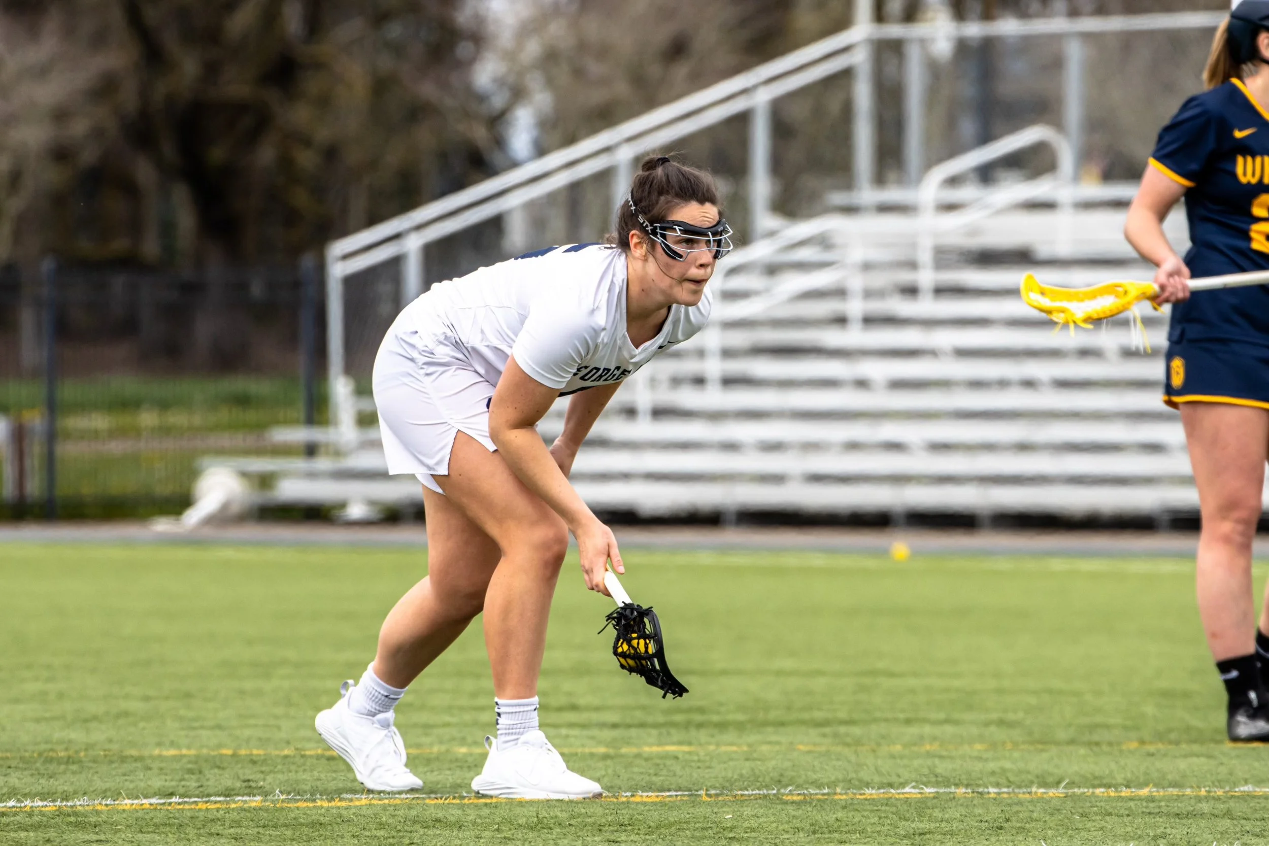 Female lacrosse player in white uniform crouching on the field with a lacrosse stick in hand, wearing goggles and looking focused for gameplay.
