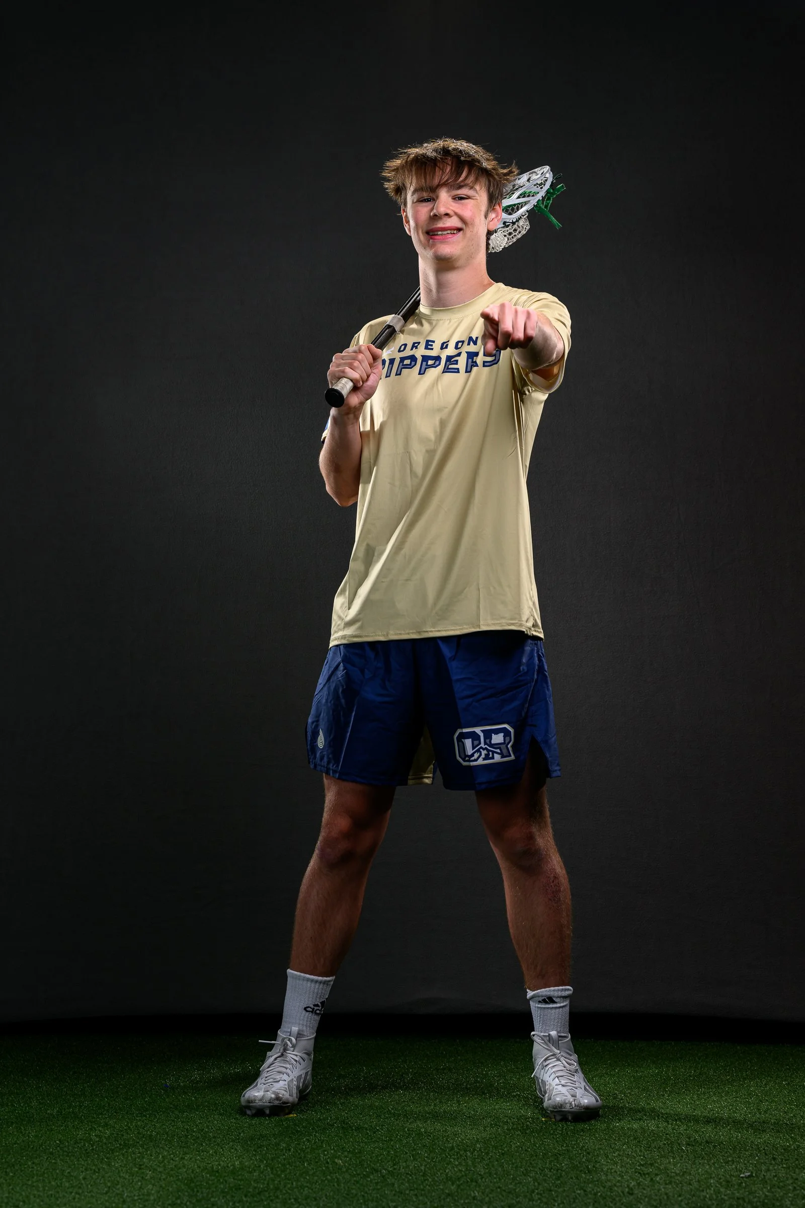 A young man wearing 'Oregon Rippers', blue shorts with logo, white athletic shoes, and white socks. He is holding a lacrosse stick over his right shoulder, smiling and pointing towards the camera against a black background.