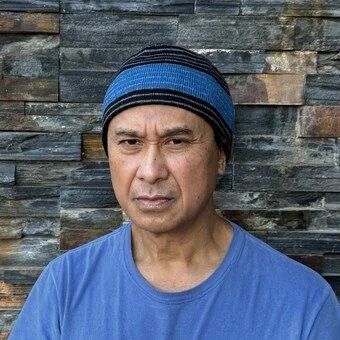 A man wearing a blue and gray striped knit beanie and a light gray long-sleeve shirt, standing in front of a stone wall.