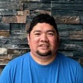 Portrait of a man with dark hair and facial hair, wearing a black T-shirt, standing in front of a multi-colored brick wall.