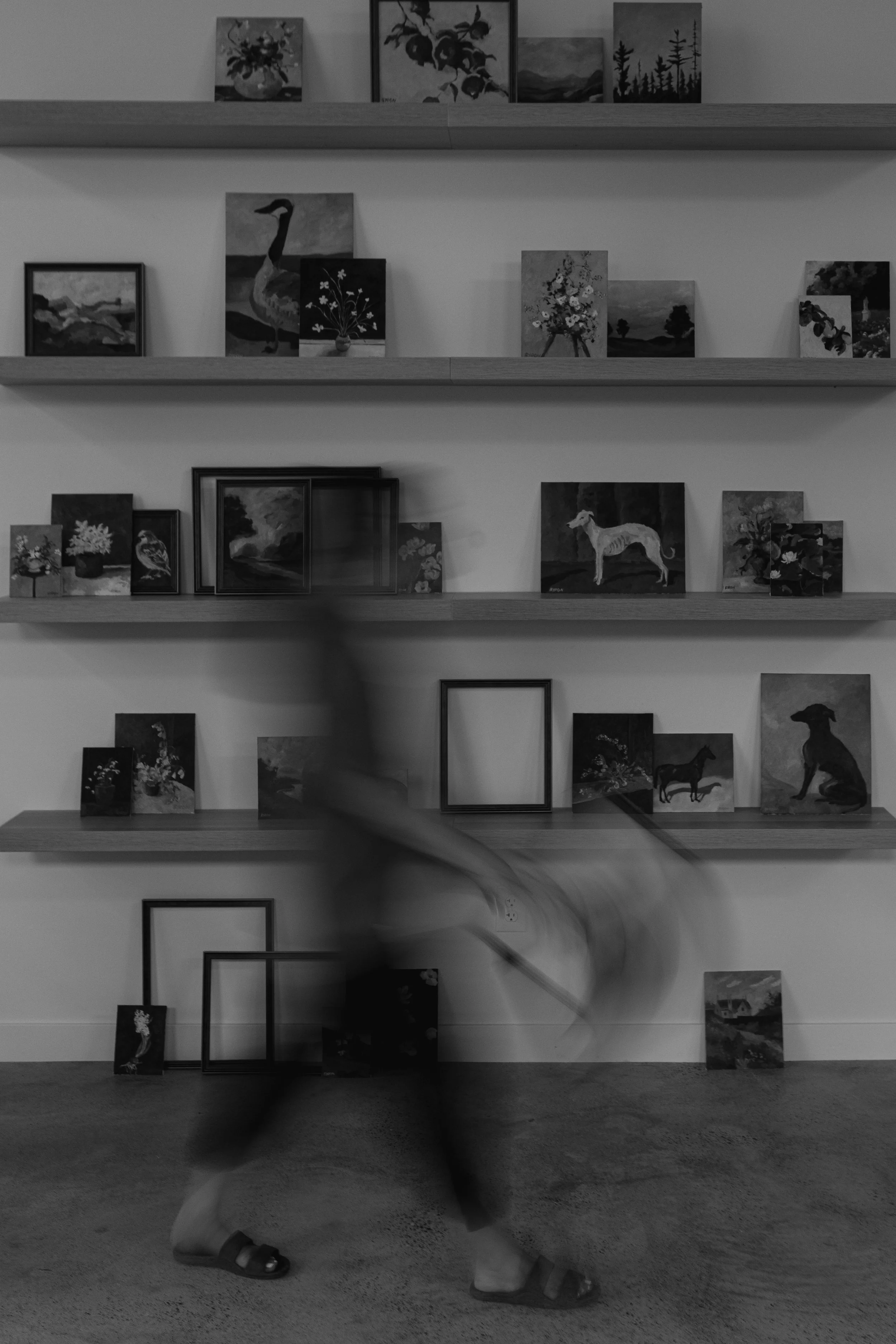A person dancing in front of a wall with shelves holding paintings and art, with some frames on the floor leaning against the wall in a black-and-white photo.