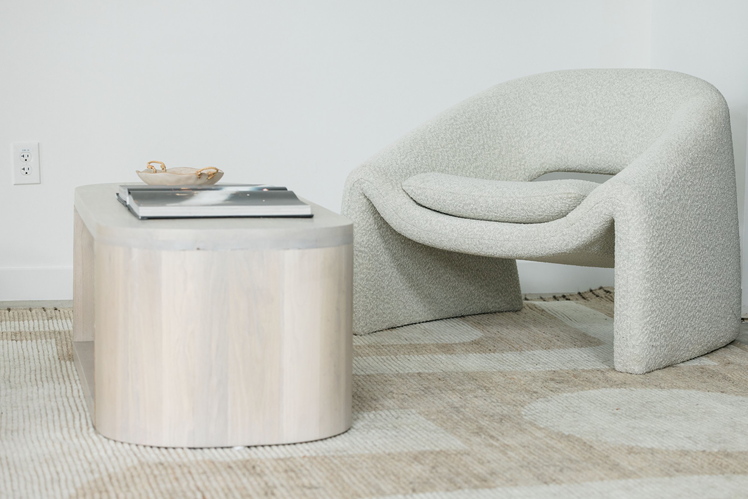 A beige upholstered armchair with a matching cushion next to a light-colored wooden side table with a glossy magazine or book on top, a small decorative bowl, and a beige and cream patterned area rug on the floor. An electrical outlet is visible on t