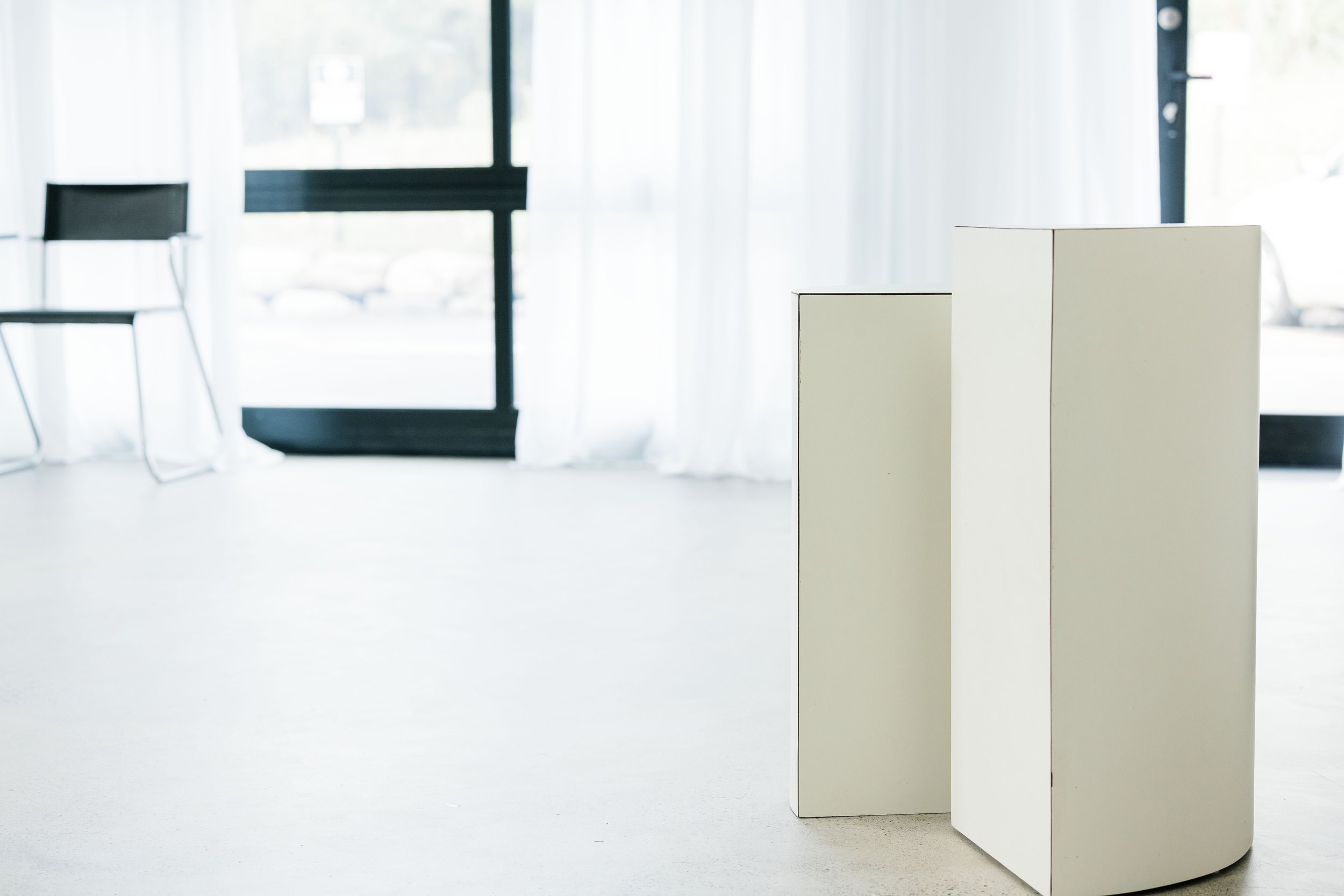 Empty room with white walls and large windows, featuring three white rectangular pedestals of varying heights.