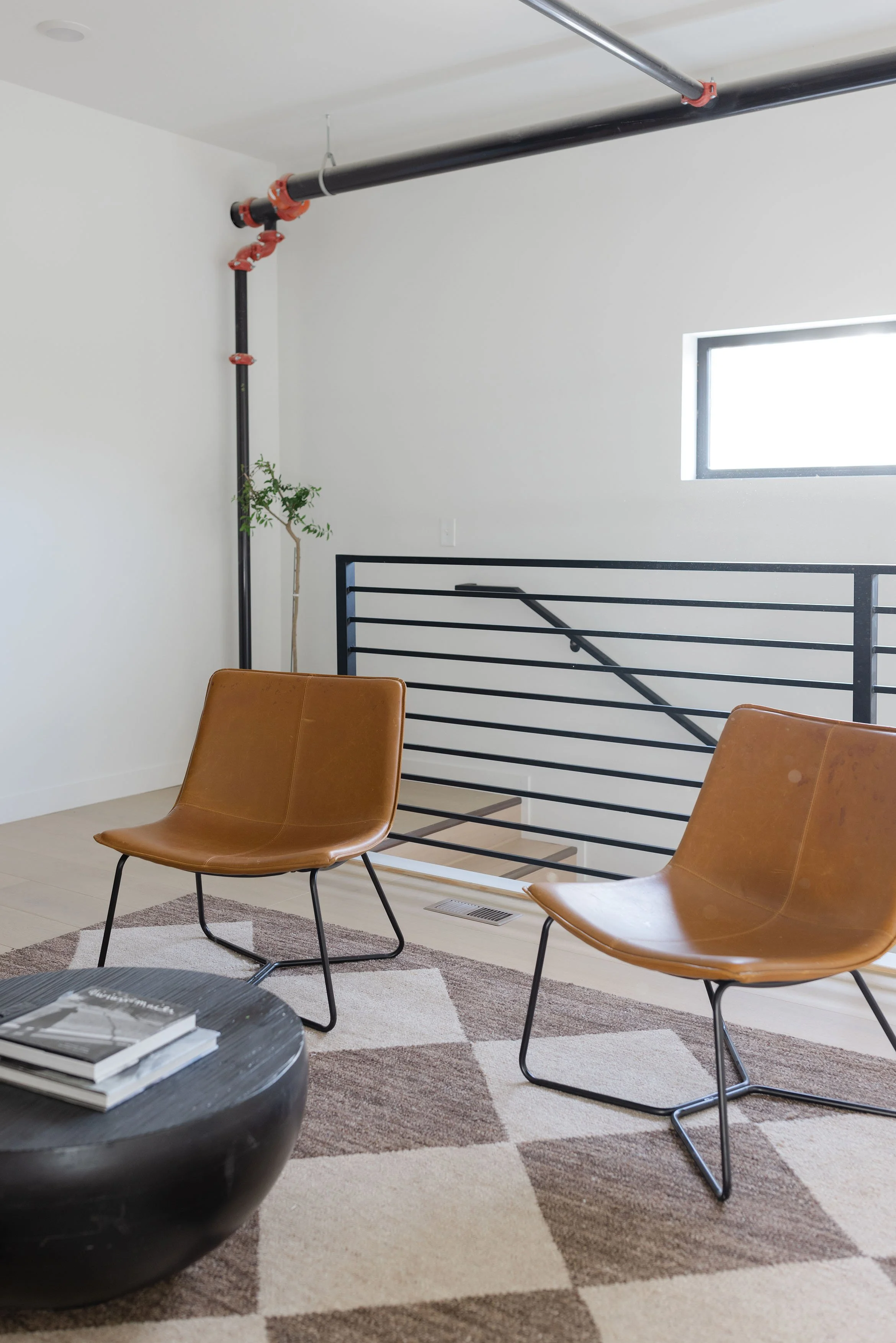 Interior of a modern living space with two tan leather chairs, a black round coffee table with a few books, a geometric patterned rug, a staircase with black railing, a small window, a potted plant, and visible industrial piping on the ceiling and corner.