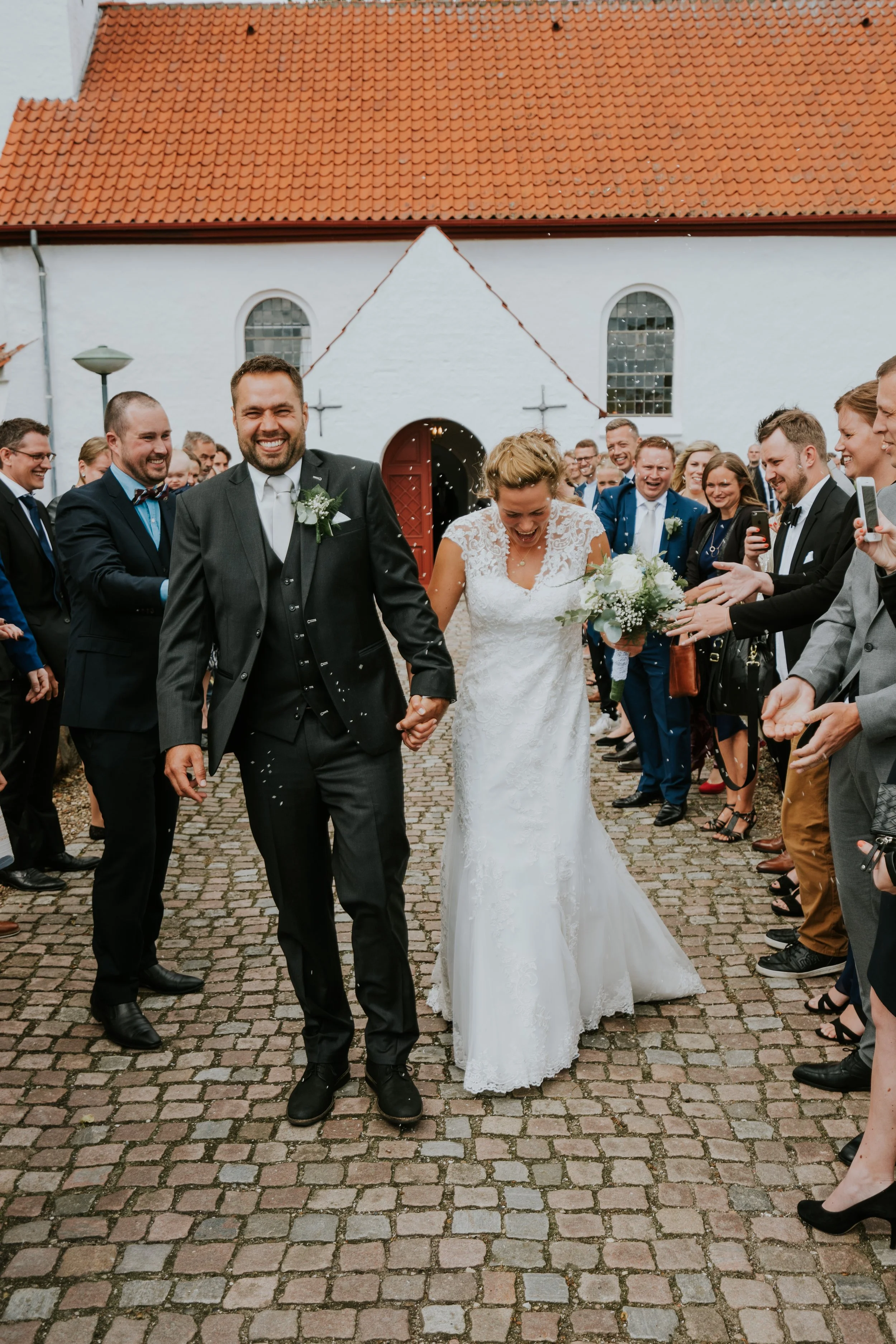En ældre brudepar går hånd i hånd ud af en kirke, omgivet af glade gæster, der kaster konfetti, i en hyggelig udendørs fest.
