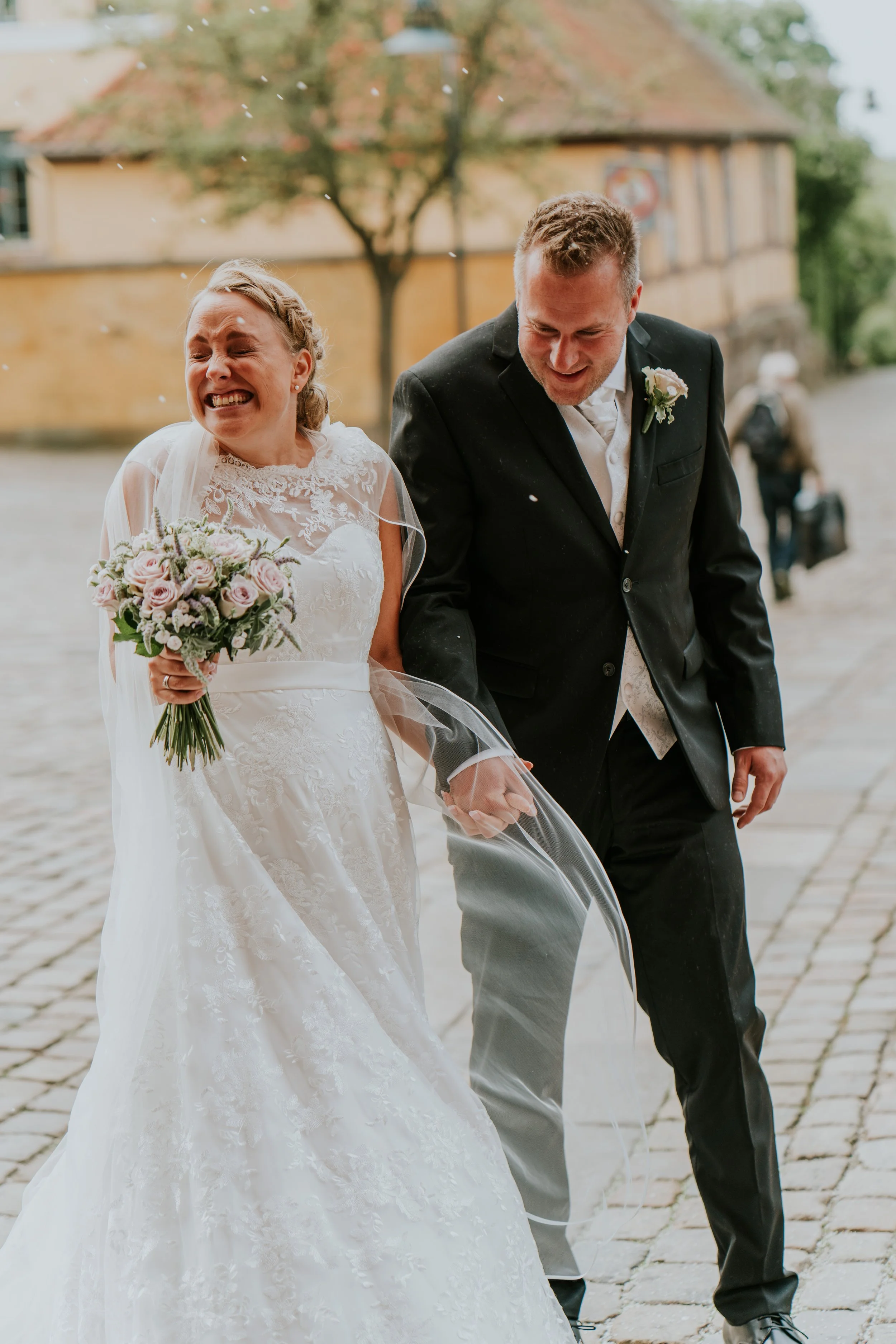 En brud og brudgom går hånd i hånd under regn, smiler og griner. Bruden bærer en hvid brudekjole og holder en buket af lyserøde og hvide blomster, mens brudgommen har jakkesæt.