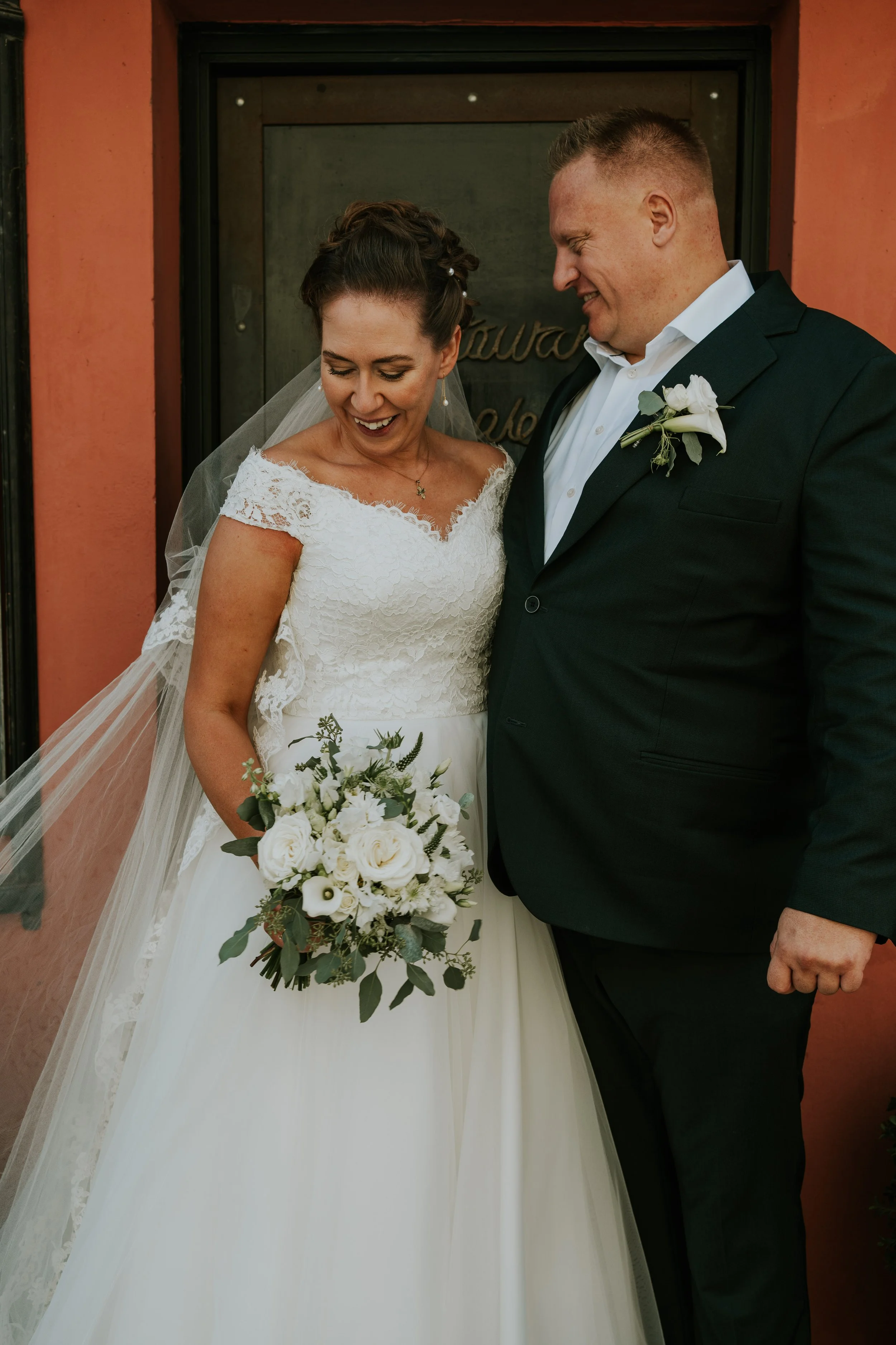 En glad brud og gommie står tæt sammen på deres bryllupsdag udenfor en bygning med en rød væg. Bruden holder en hvid blomsterbuket, og de er begge klædt i formelt tøj.