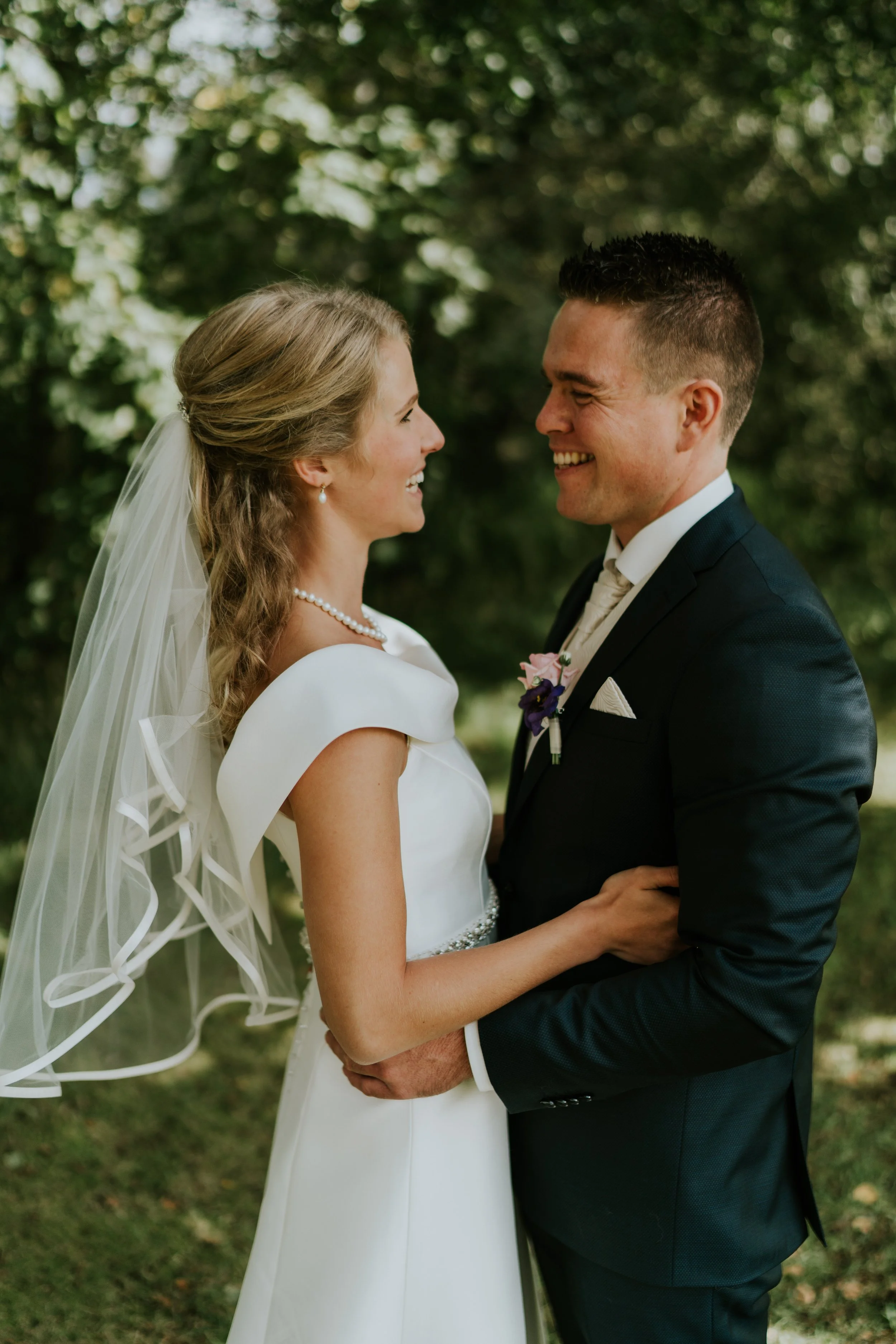 To brudepar står tæt sammen og smiler i en udendørs skov, iført bryllupsudstyr.