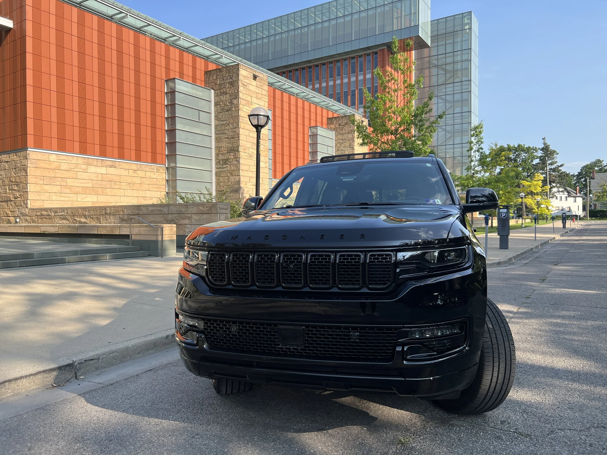 Black Wagoneer SUV parked on a city street with modern red and glass buildings in the background, trees, and a clear blue sky.