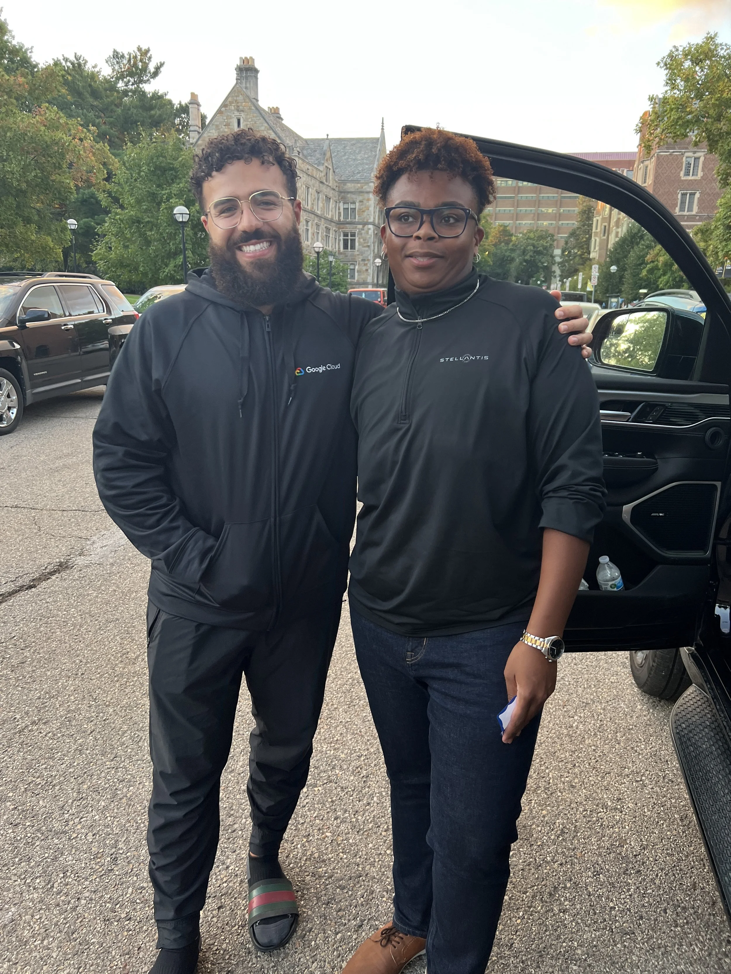 Two people standing outside near cars and a historic building, smiling at the camera. One is a man with a beard, glasses, and a black jacket with Google Cloud logo. The other is a woman with short hair, glasses, and a black jacket with Stellaantis lo