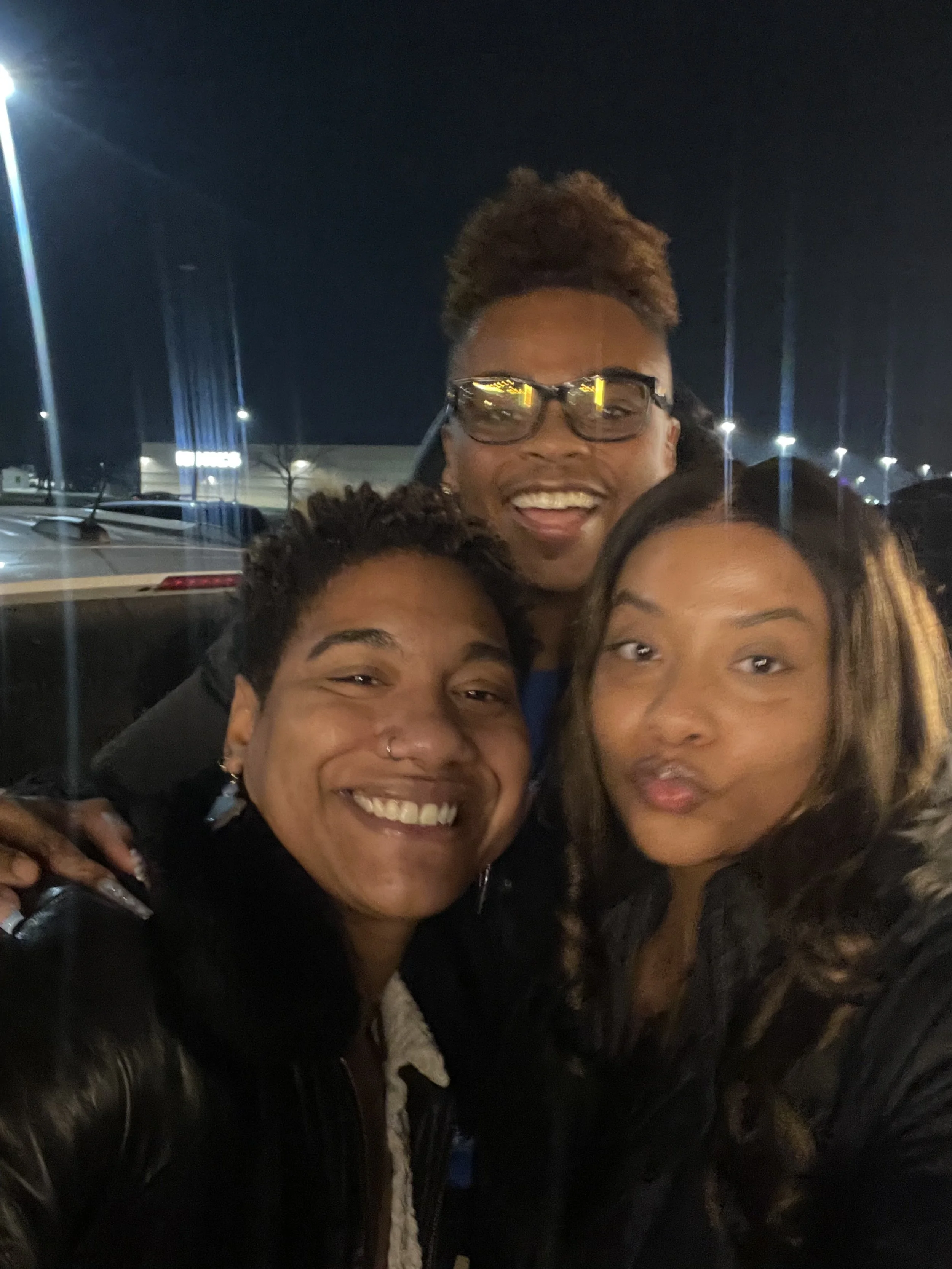 Smiling group of three women and one man taking a selfie outside at night, with dim street lights and parked cars in the background.