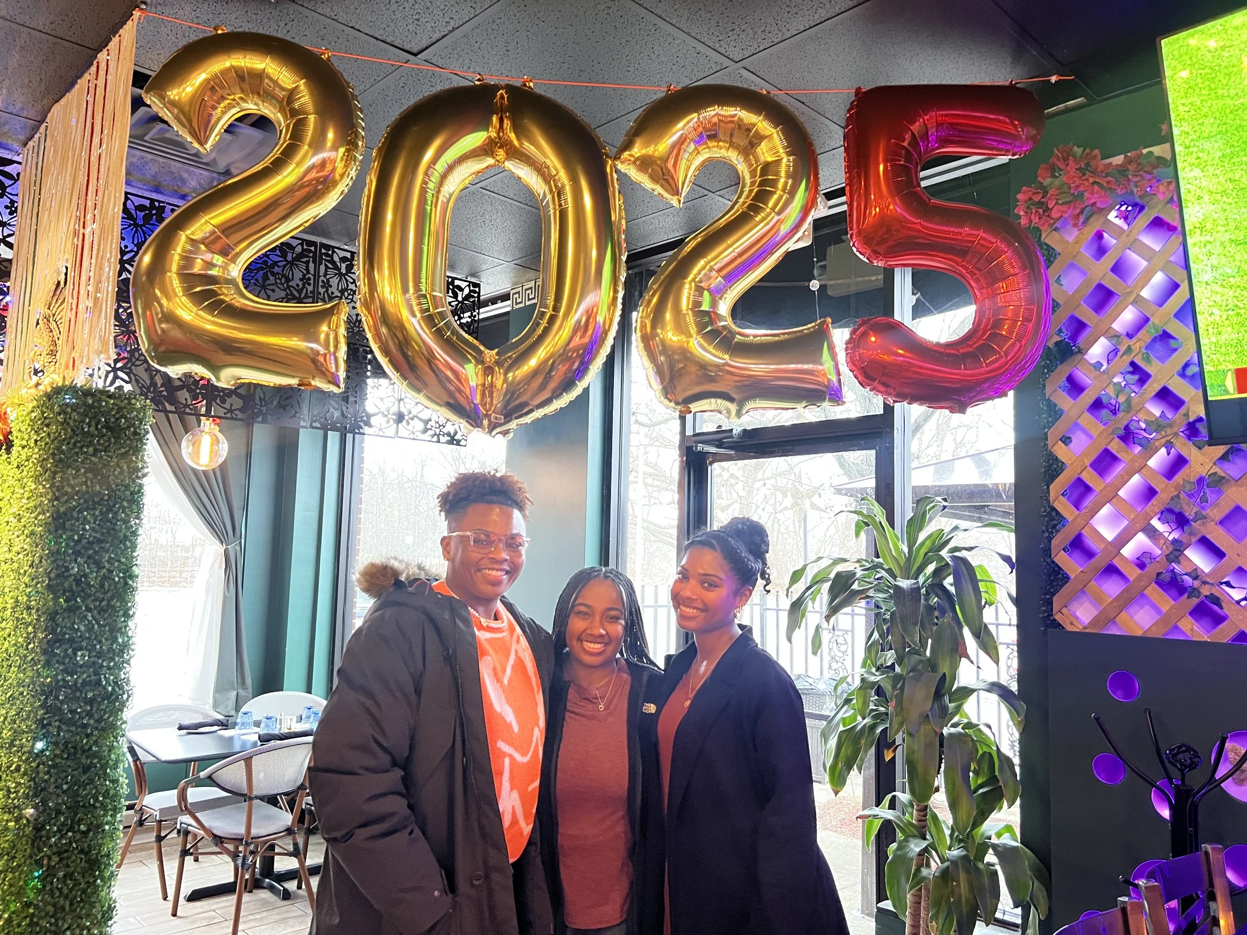 Three smiling women standing inside a room decorated with large gold and red balloons spelling out 2023, with a window and potted plants in the background.
