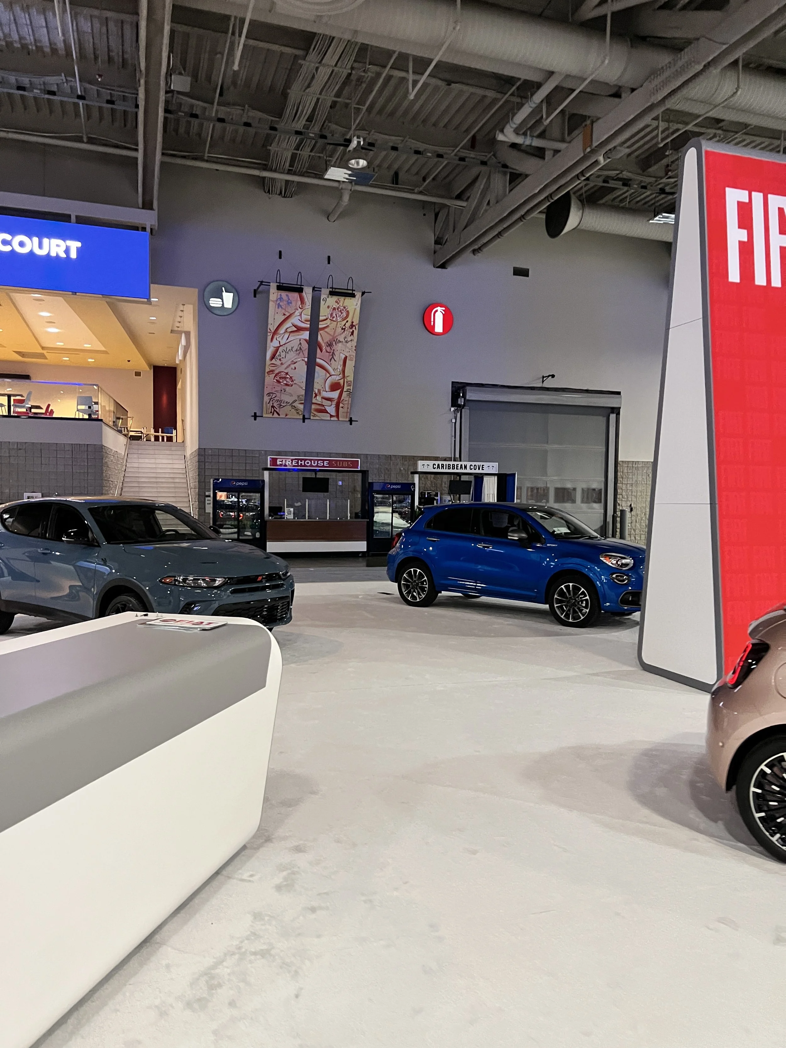 Indoor car dealership with various cars, a large red sign, and signage for fire safety and a subway station in the background.