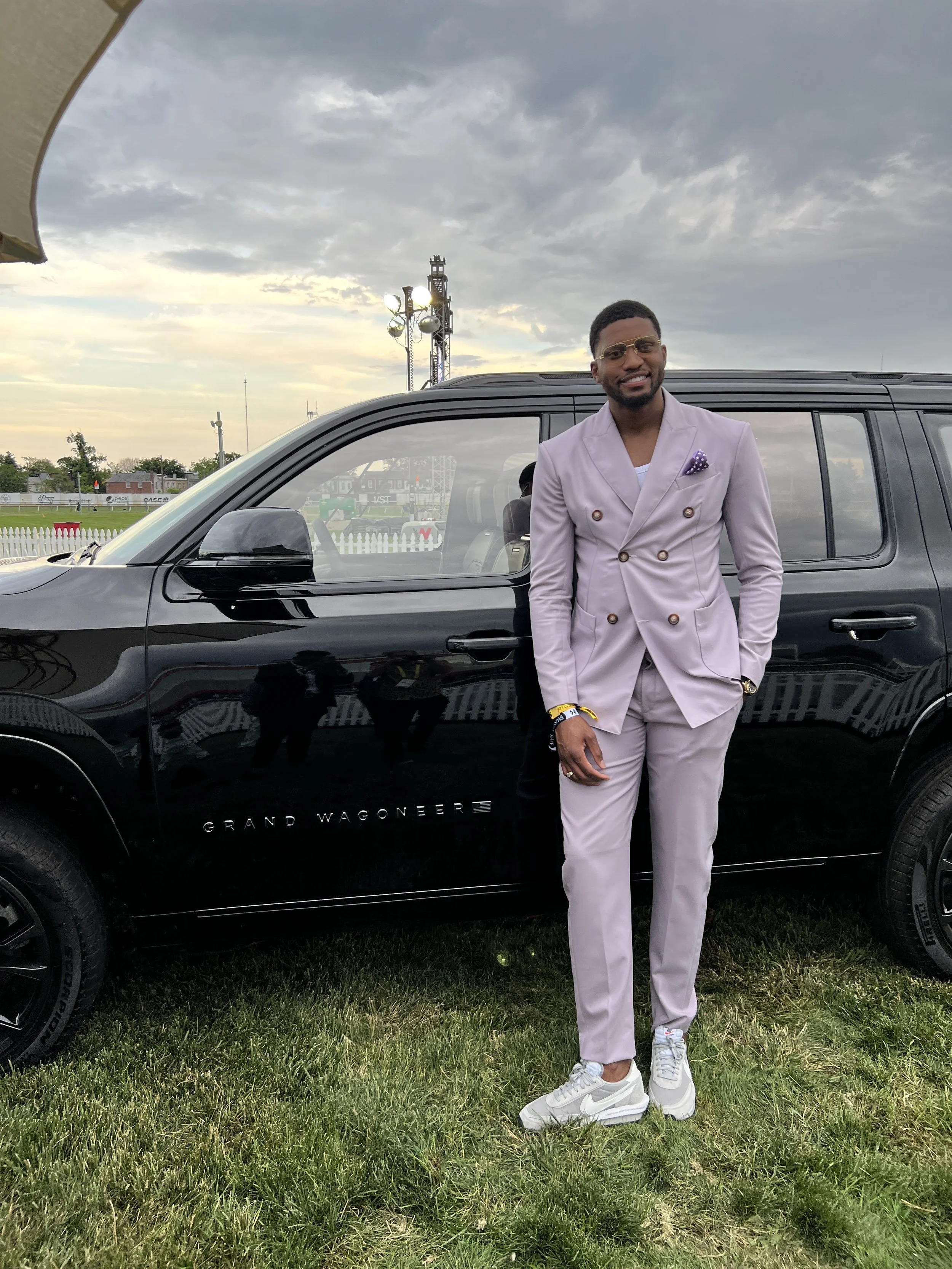 A man in a light purple suit stands outside next to a black Grand Wagoneer vehicle on a grassy field. He's wearing white sneakers and has sunglasses on, with a cloudy sky and some event lighting structures in the background.