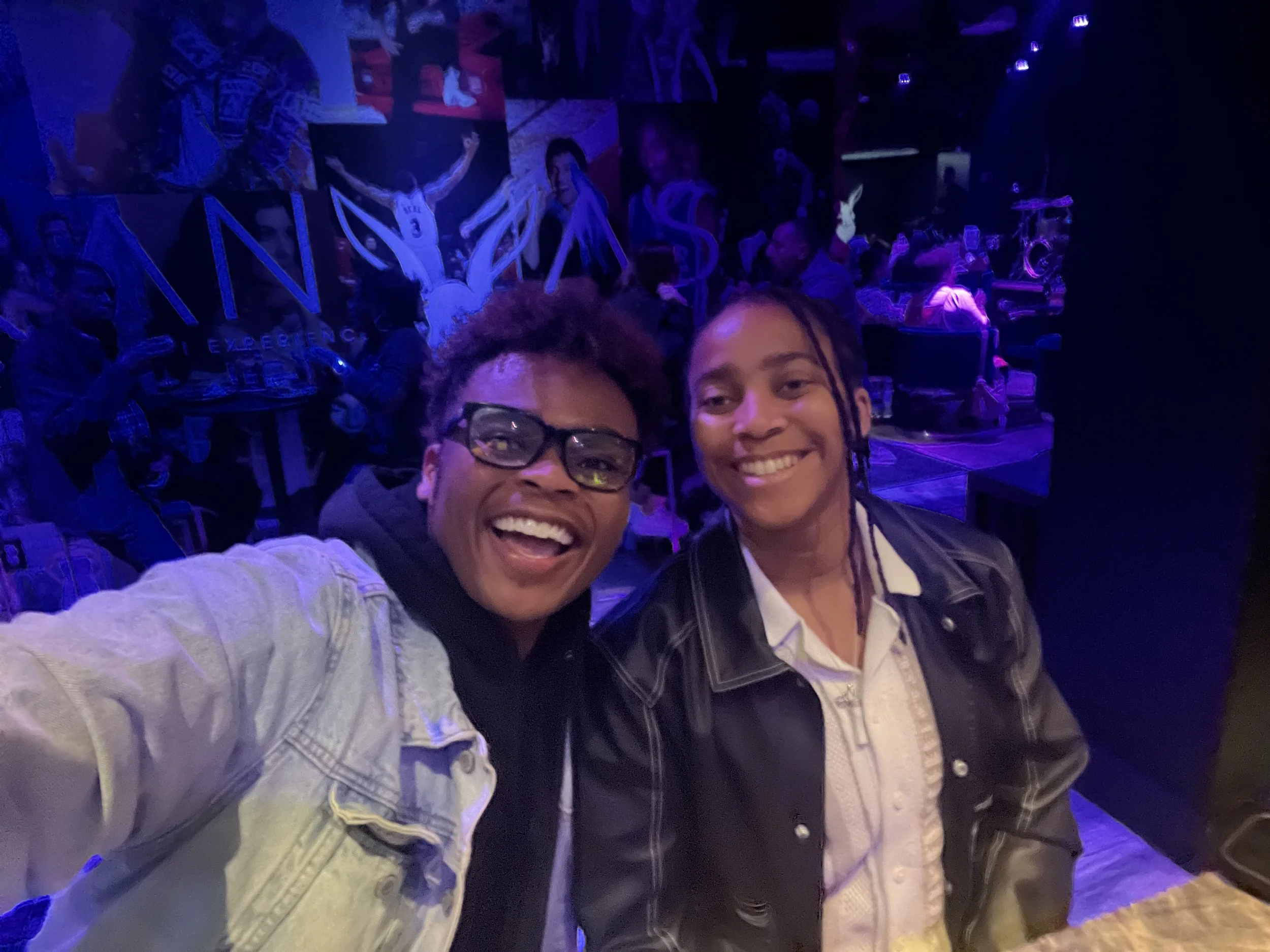 Two smiling individuals taking a selfie in a dimly lit, colorful environment, possibly a bar or nightclub, with a group of people and vibrant signs in the background.