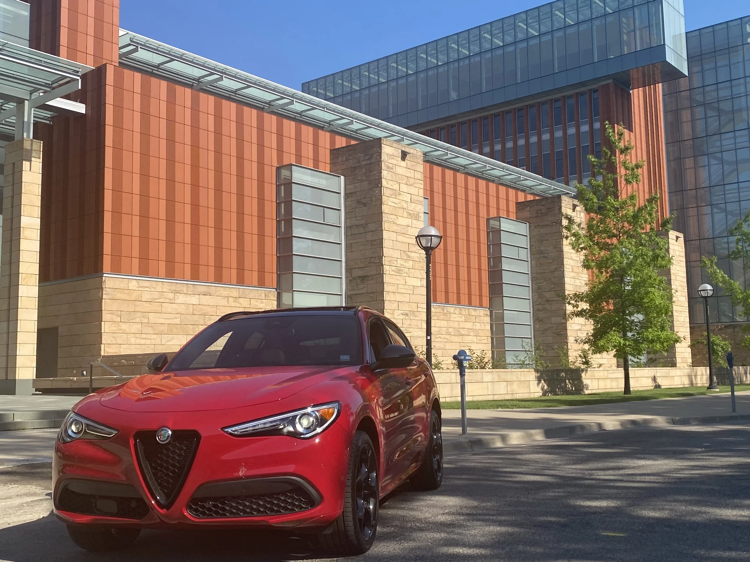 Red Alfa Romeo parked on city street in front of modern brick and glass building with trees and streetlamps.