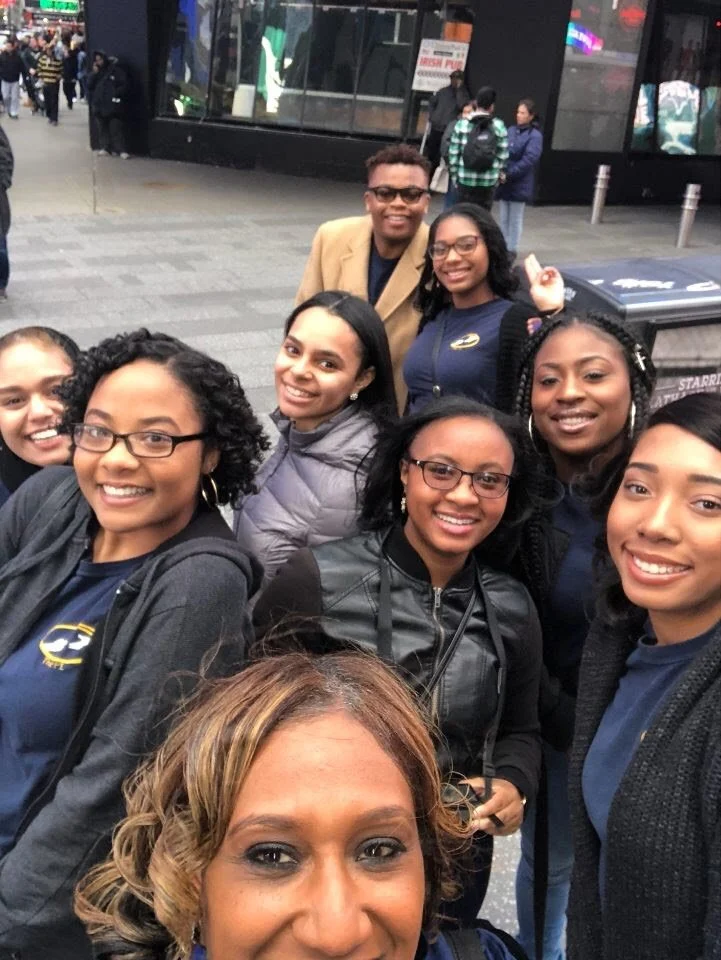 A group of nine smiling people taking a selfie outside a building, with some wearing matching navy shirts. The group includes one man and eight women of various ages and ethnicities.