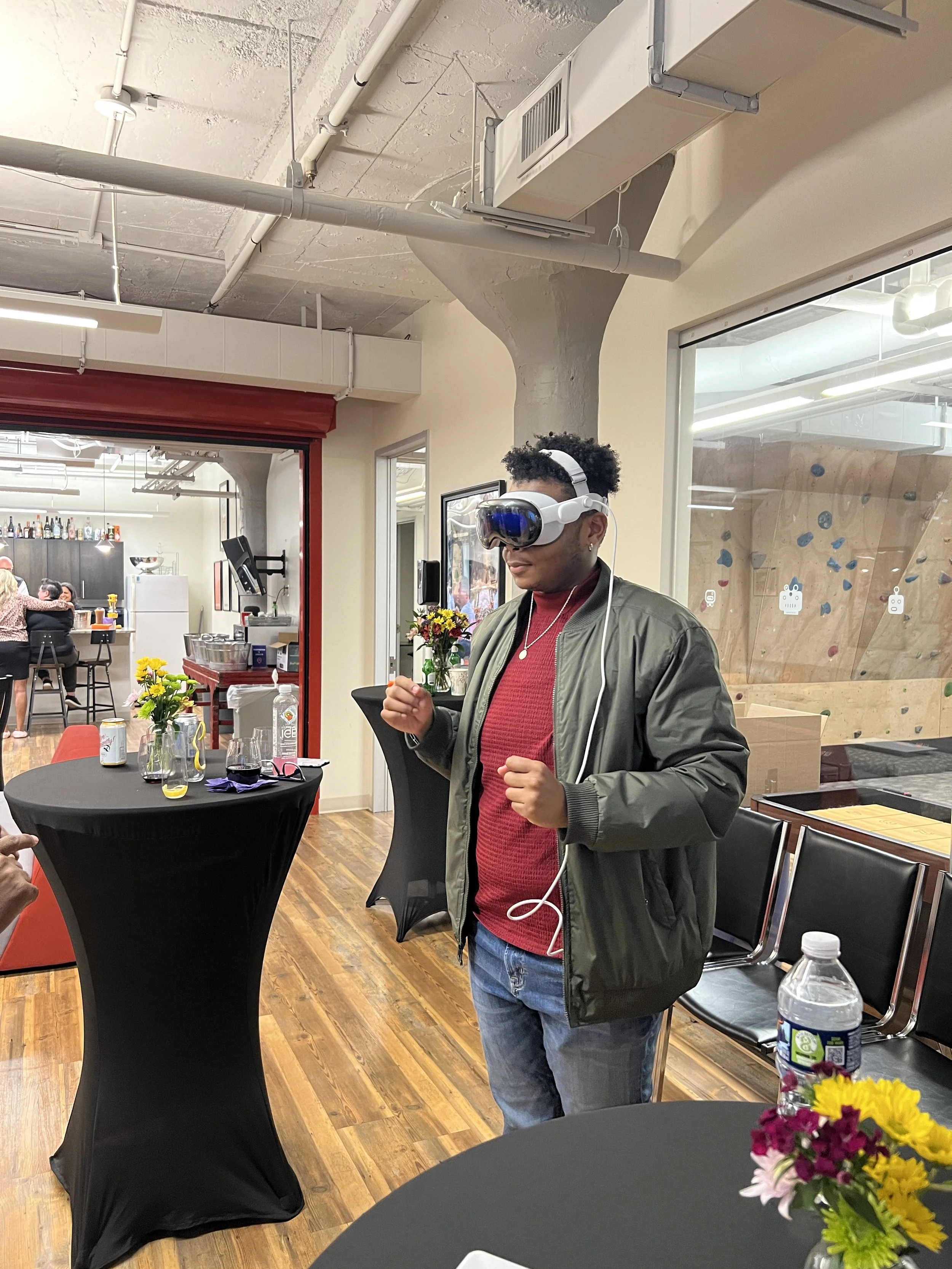 A young person wearing a VR headset and headphones, standing indoors at a social event or gathering, with tables decorated with flowers, and people in the background.