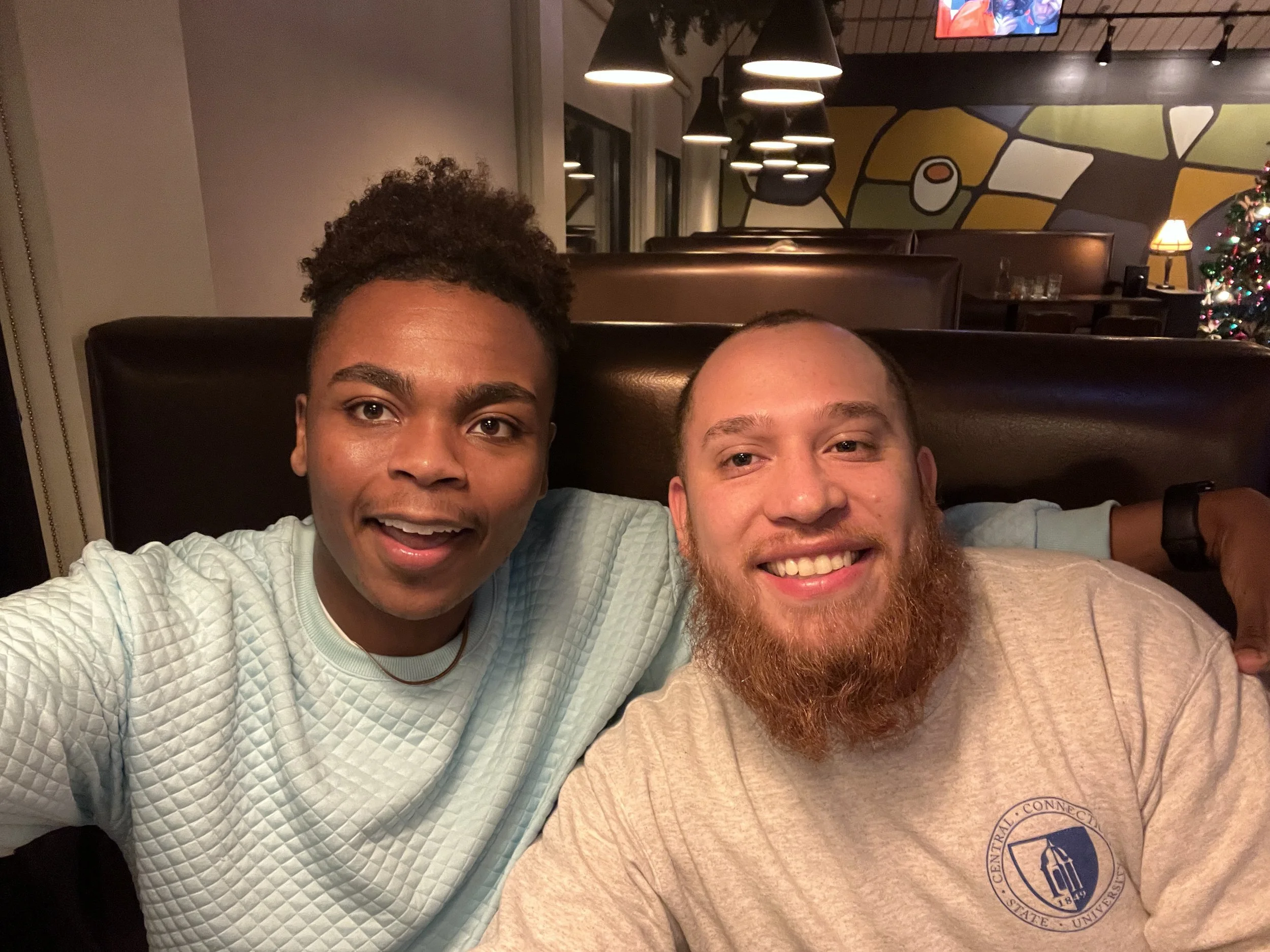 Two men smiling and taking a selfie inside a restaurant, with a decorated Christmas tree and colorful wall art in the background.