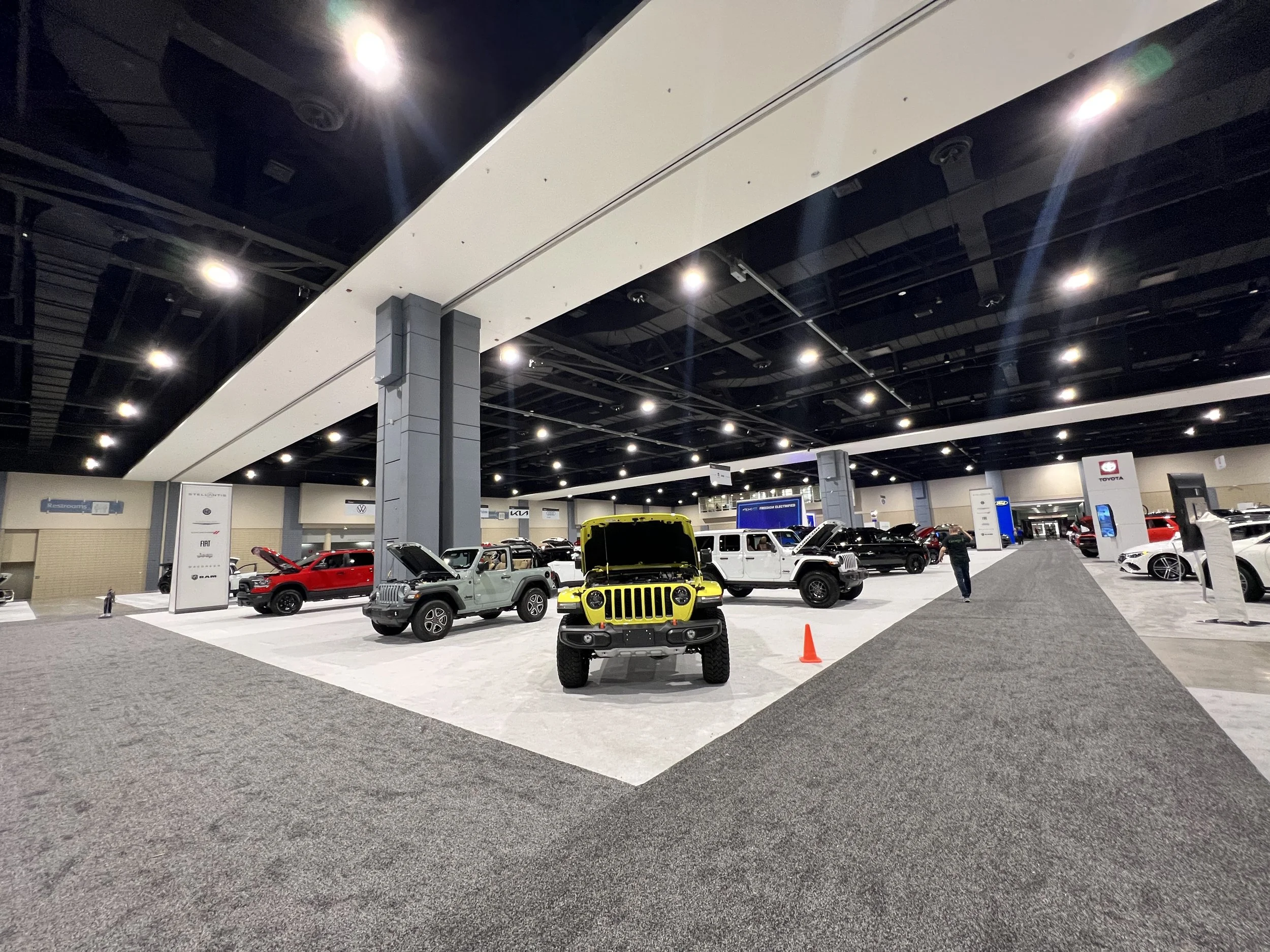 Indoor car showroom with various new cars, including a bright yellow Jeep with open hood, and a gray Jeep, with some signs and a person walking.