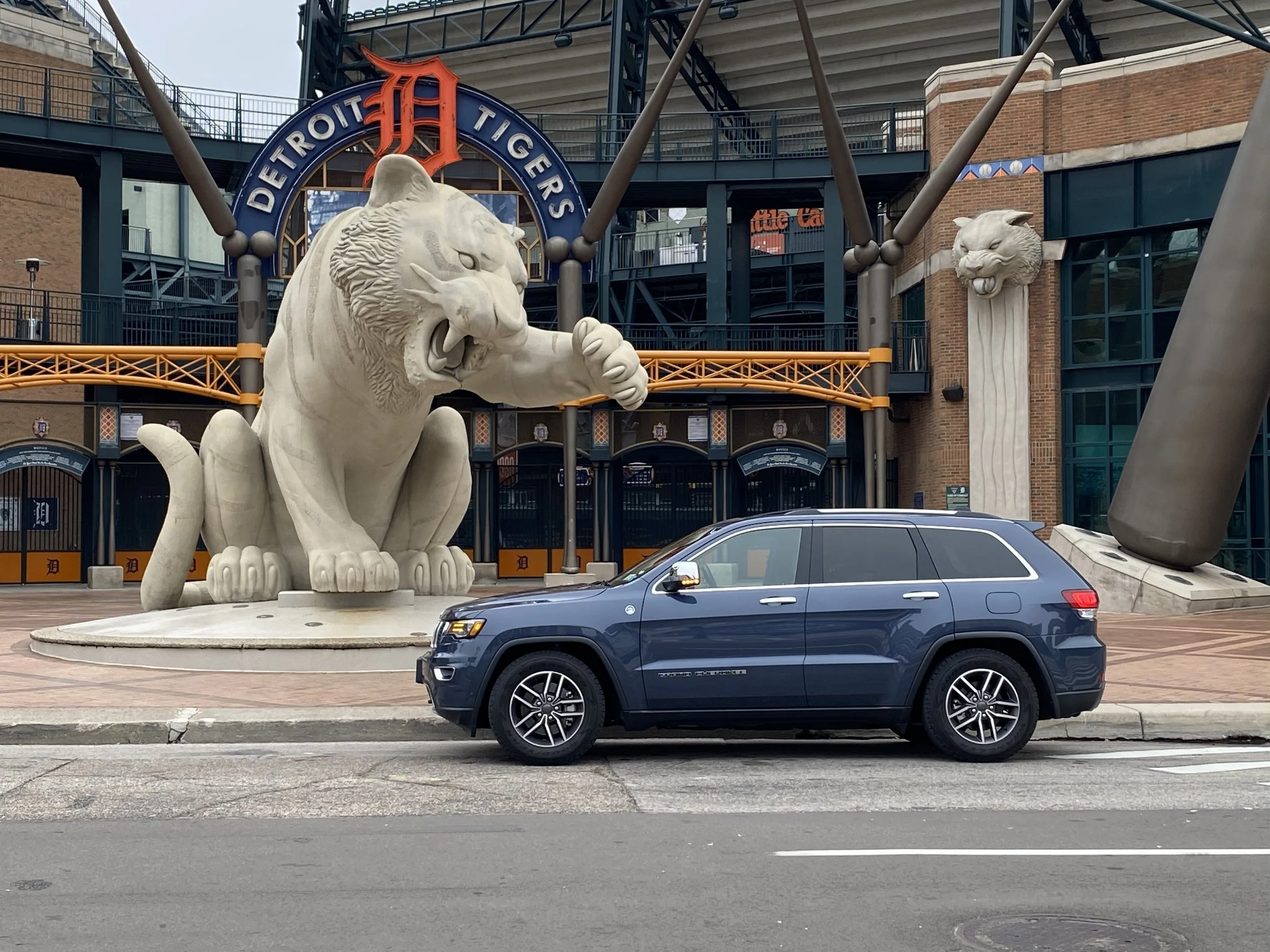 Detroit Tigers stadium entrance with large lion statues, a black SUV parked in front, and a baseball bat sculpture.