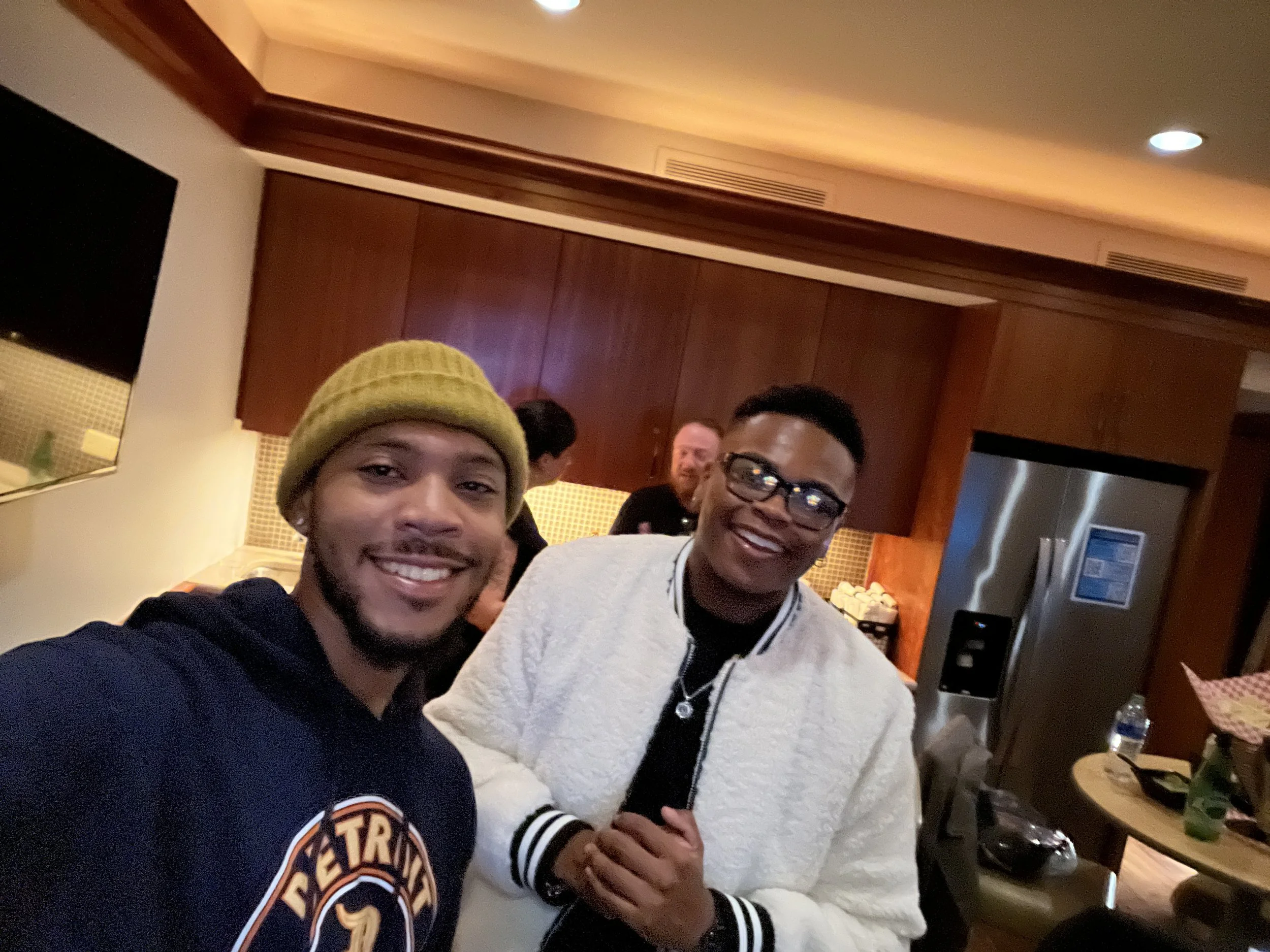 Two young men smiling indoors, taking a selfie together at a social gathering, with a woman in the background and kitchen area visible.