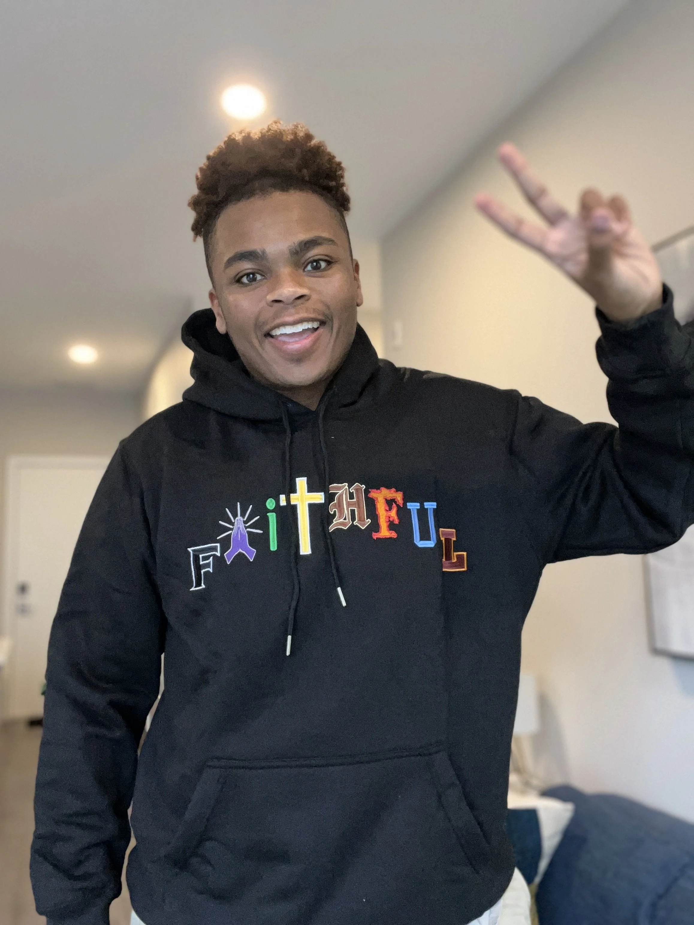 A young man with short curly hair smiling and making a peace sign with his right hand, wearing a black hoodie with colorful embroidered letters spelling 'FFTHFUL' on the front.