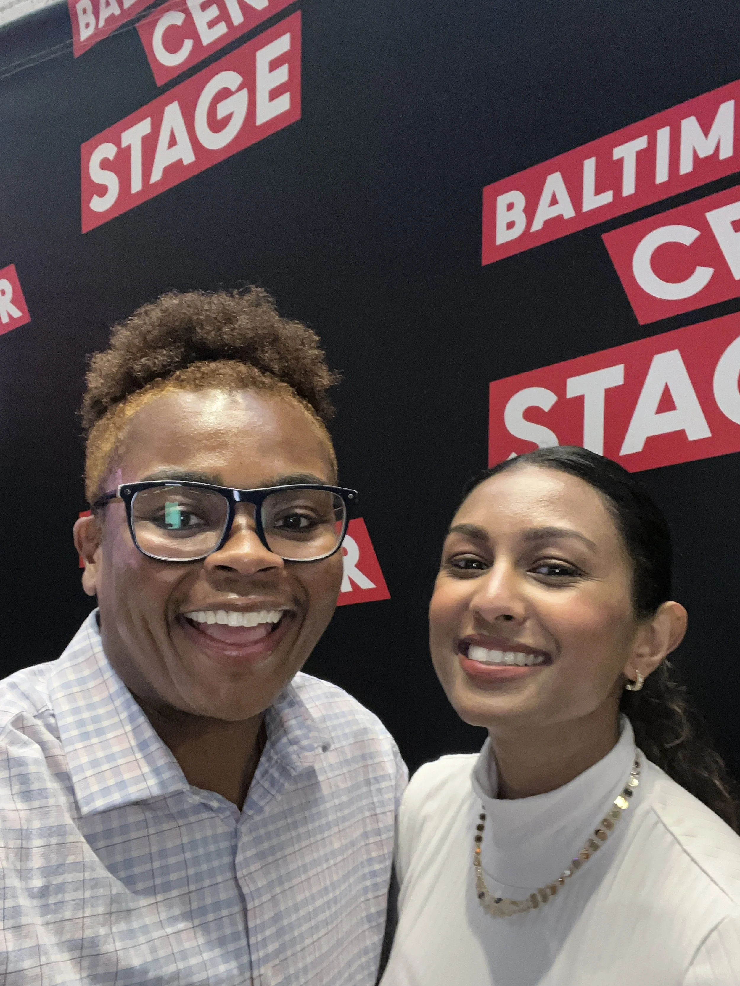 Two smiling women taking a selfie in front of a black background with red and white "Baltimore Center Stage" logos.