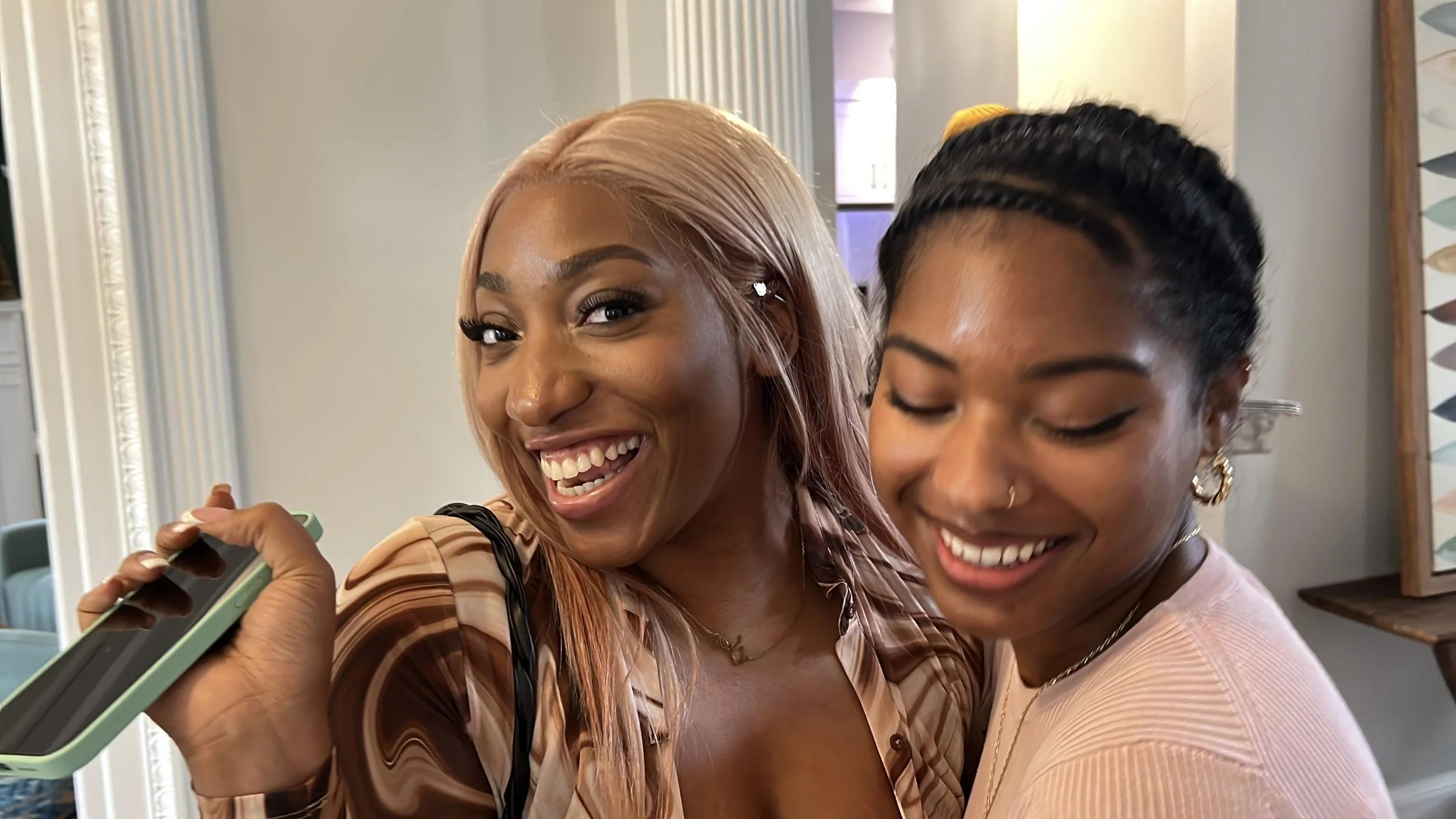 Two women smiling and hugging, one with blonde hair holding a smartphone, in a warmly lit indoor setting.