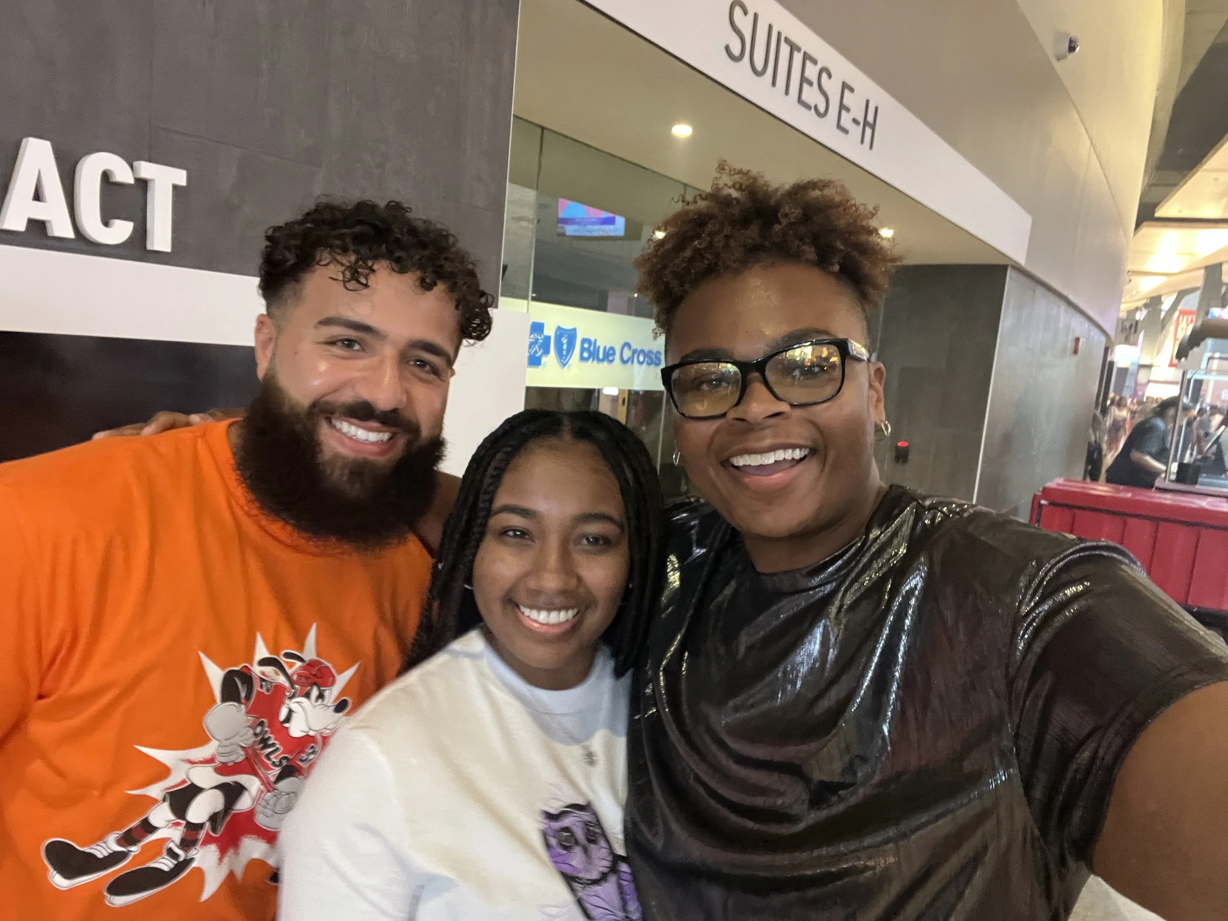 Three smiling people taking a selfie in an indoor location, with signs and a Blue Cross logo visible in the background.