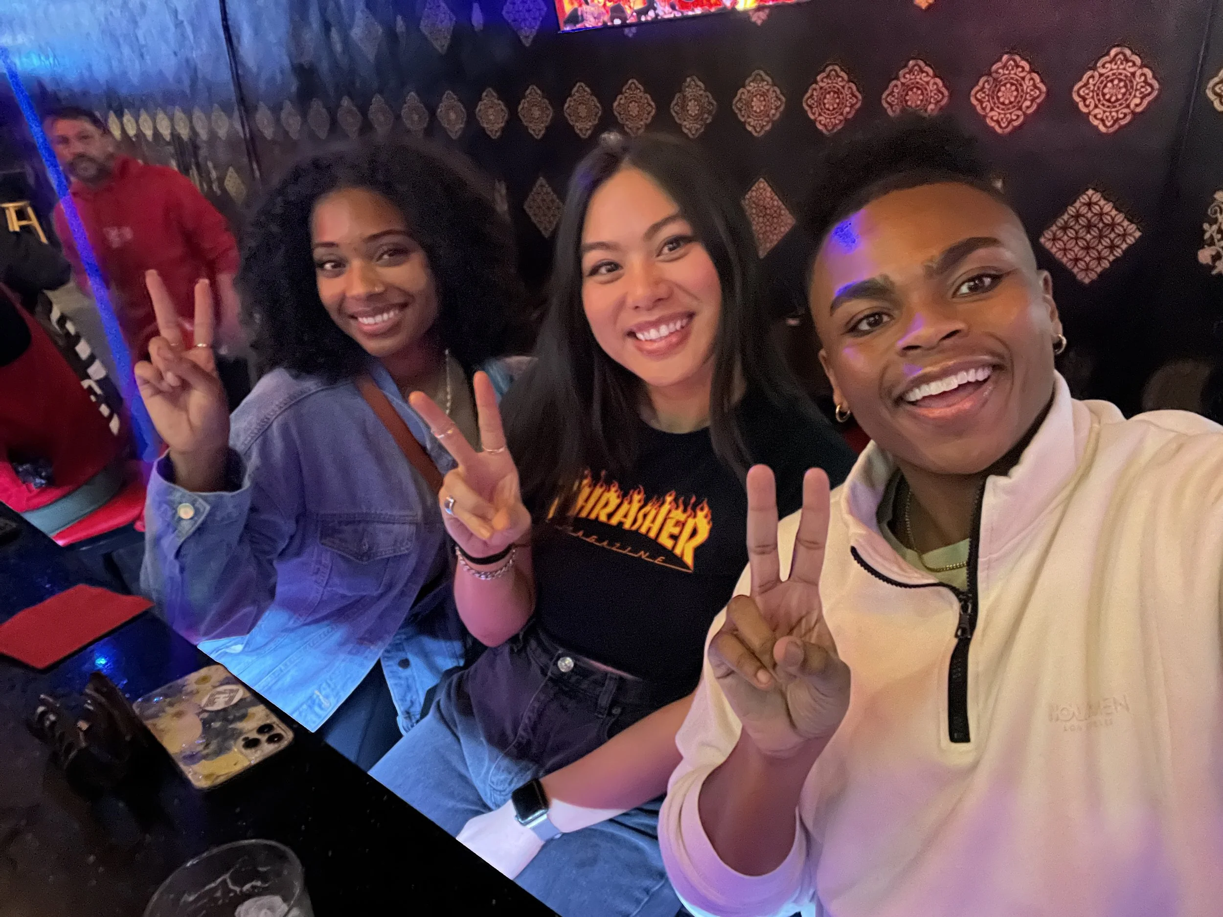 Three women sitting at a table, smiling, and making peace signs. The woman on the left has curly hair and a denim jacket, the woman in the middle has straight black hair and a black Thrasher shirt, and the woman on the right has short hair, earrings,