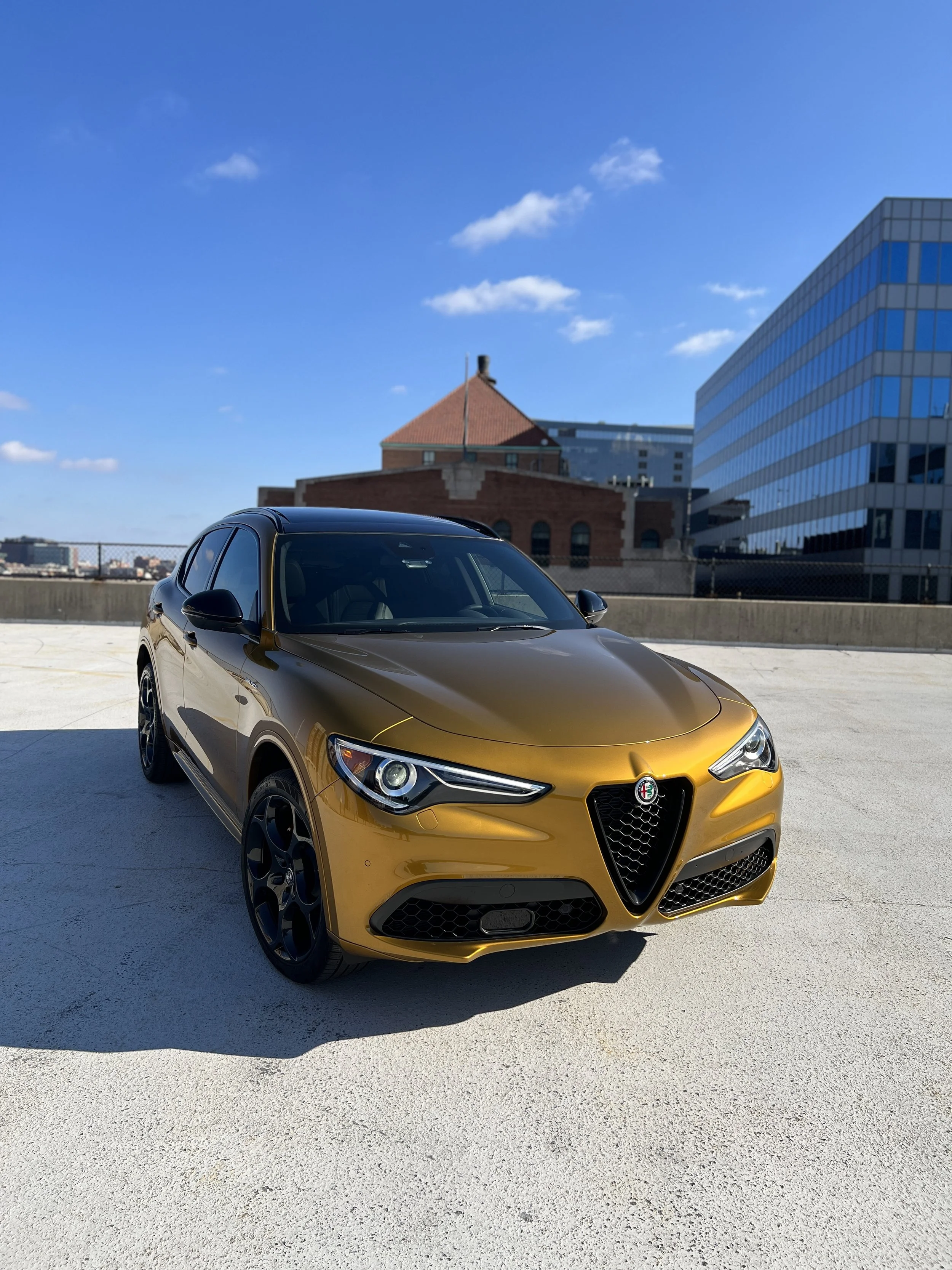 A gold-colored Alfa Romeo SUV parked on a rooftop parking lot with a cityscape in the background, including a brick building with a triangular roof and a modern glass building under a blue sky with a few clouds.
