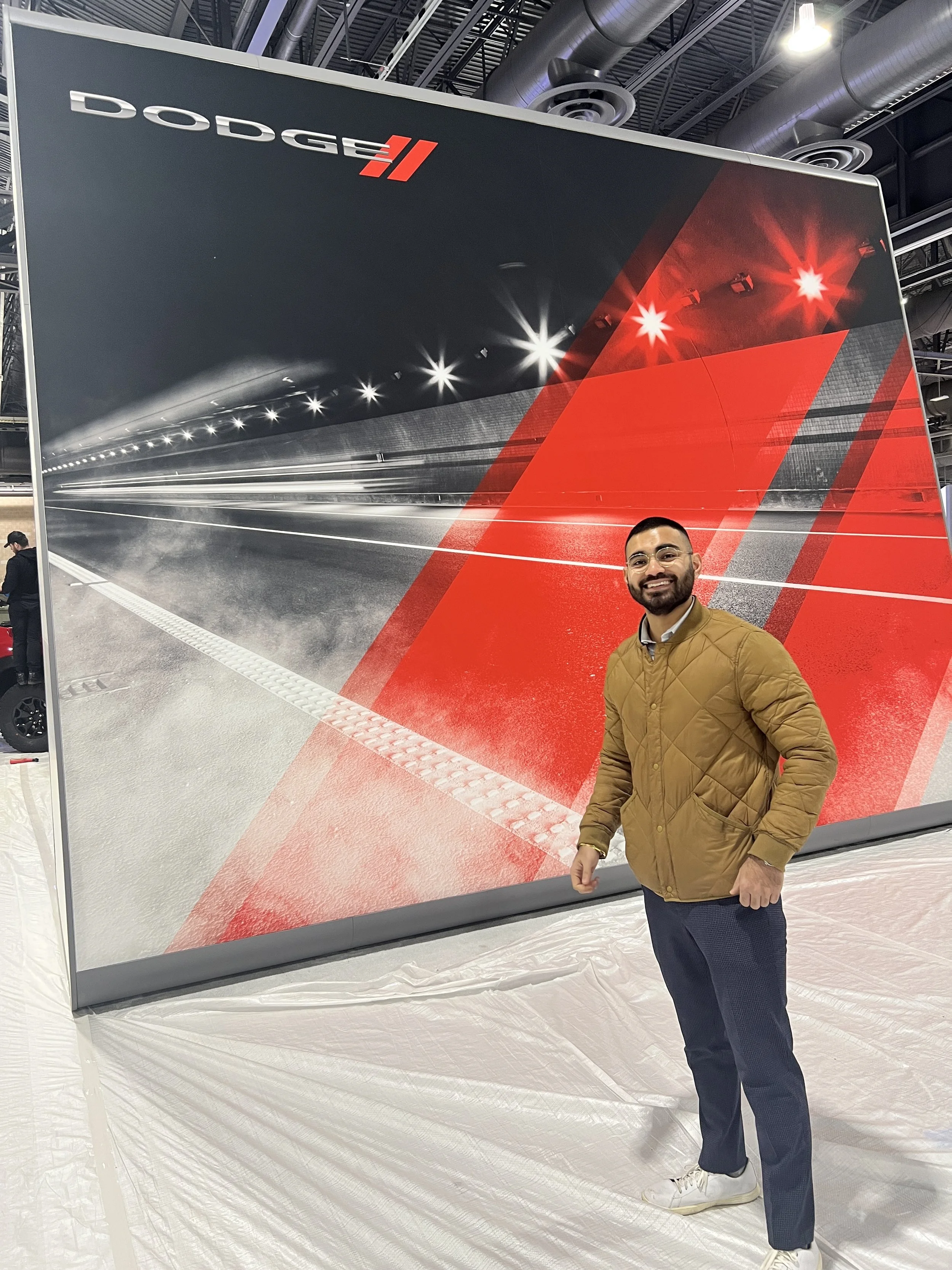 A man with glasses and a beard smiling in front of a large Dodge II advertisement at an auto show, featuring a stylized depiction of a race track with red and white lighting effects.
