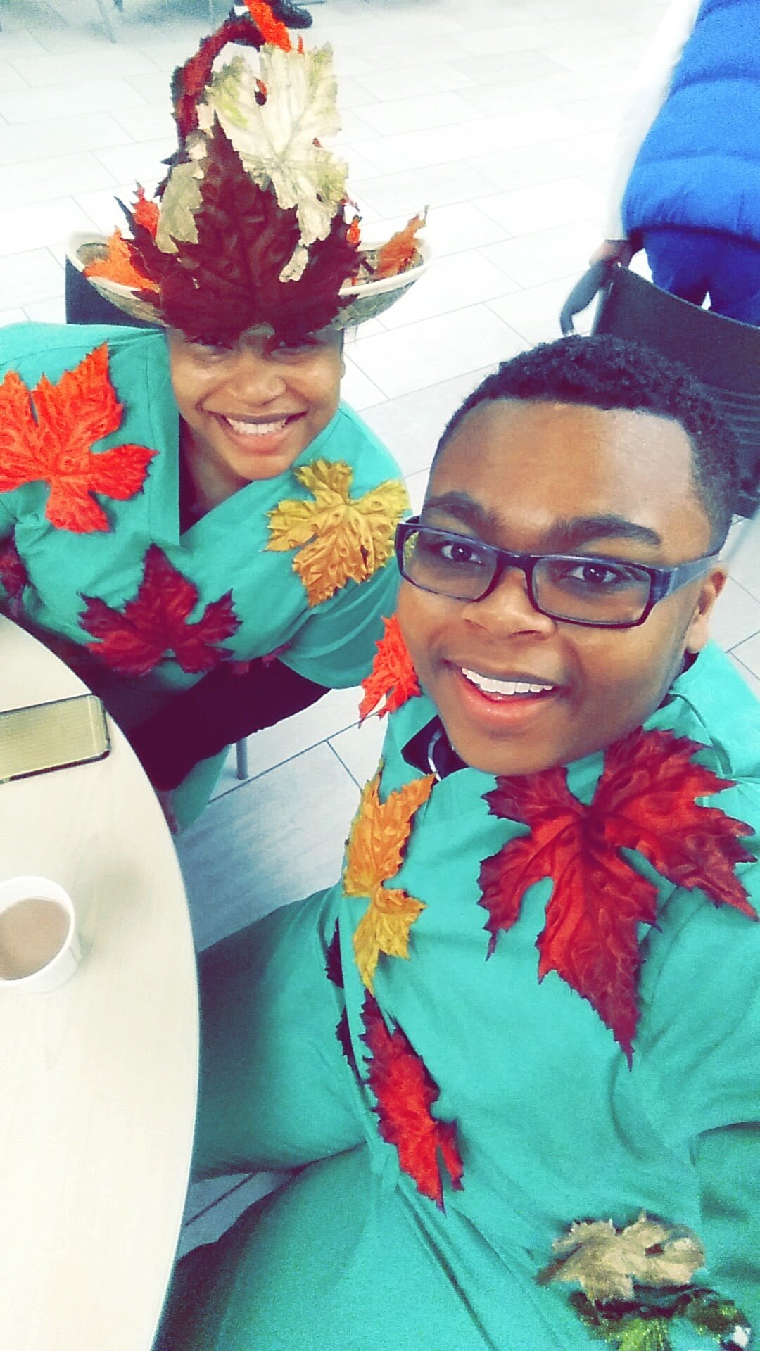Two people smiling, wearing turquoise outfits decorated with bright autumn leaves, with a woman wearing a hat made of fall leaves, standing indoors in a tiled room.