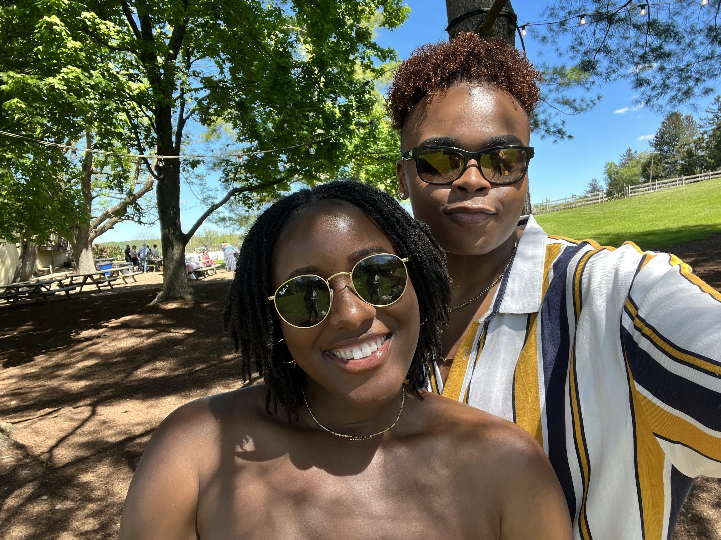 Two women smiling and taking a selfie outdoors under trees with green leaves on a sunny day, with a grassy field and a wooden fence in the background.