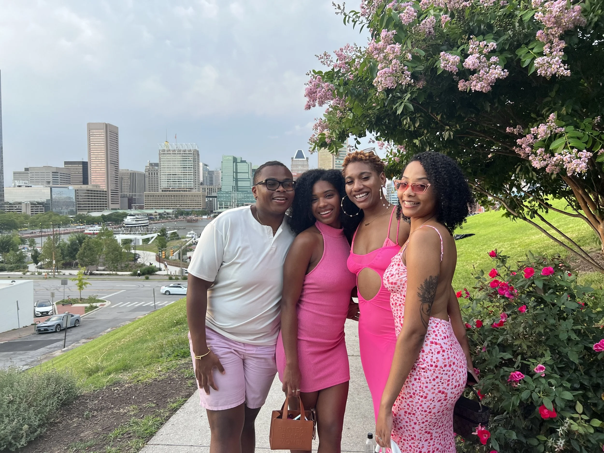 Four women standing together outdoors on a sidewalk near a bush with pink flowers, with a city skyline and river in the background. Two women are wearing pink dresses, one in a bright pink dress with cutouts, another in a pink patterned dress, and a 