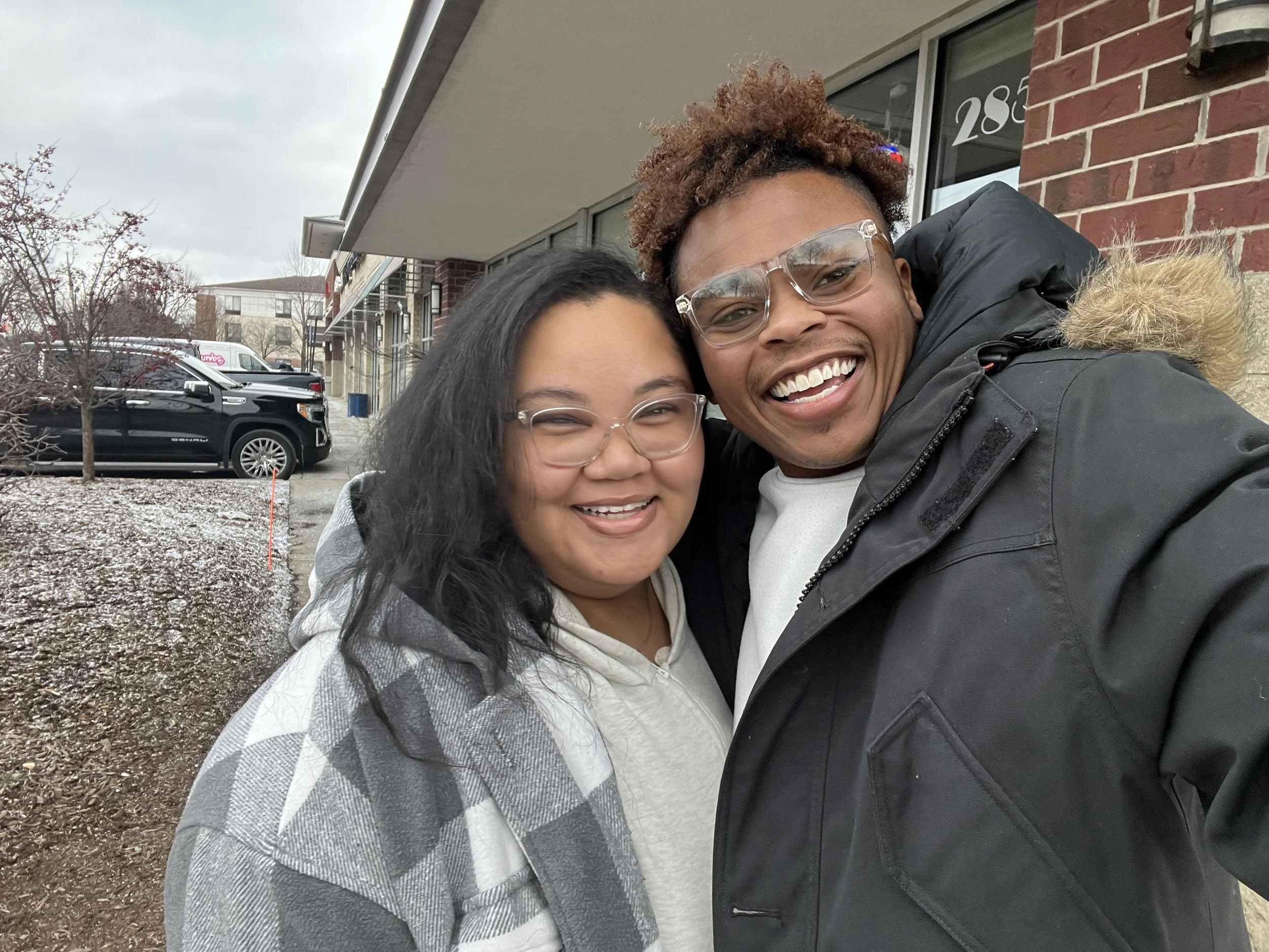 Two smiling friends taking a selfie outside a building on a cloudy day.