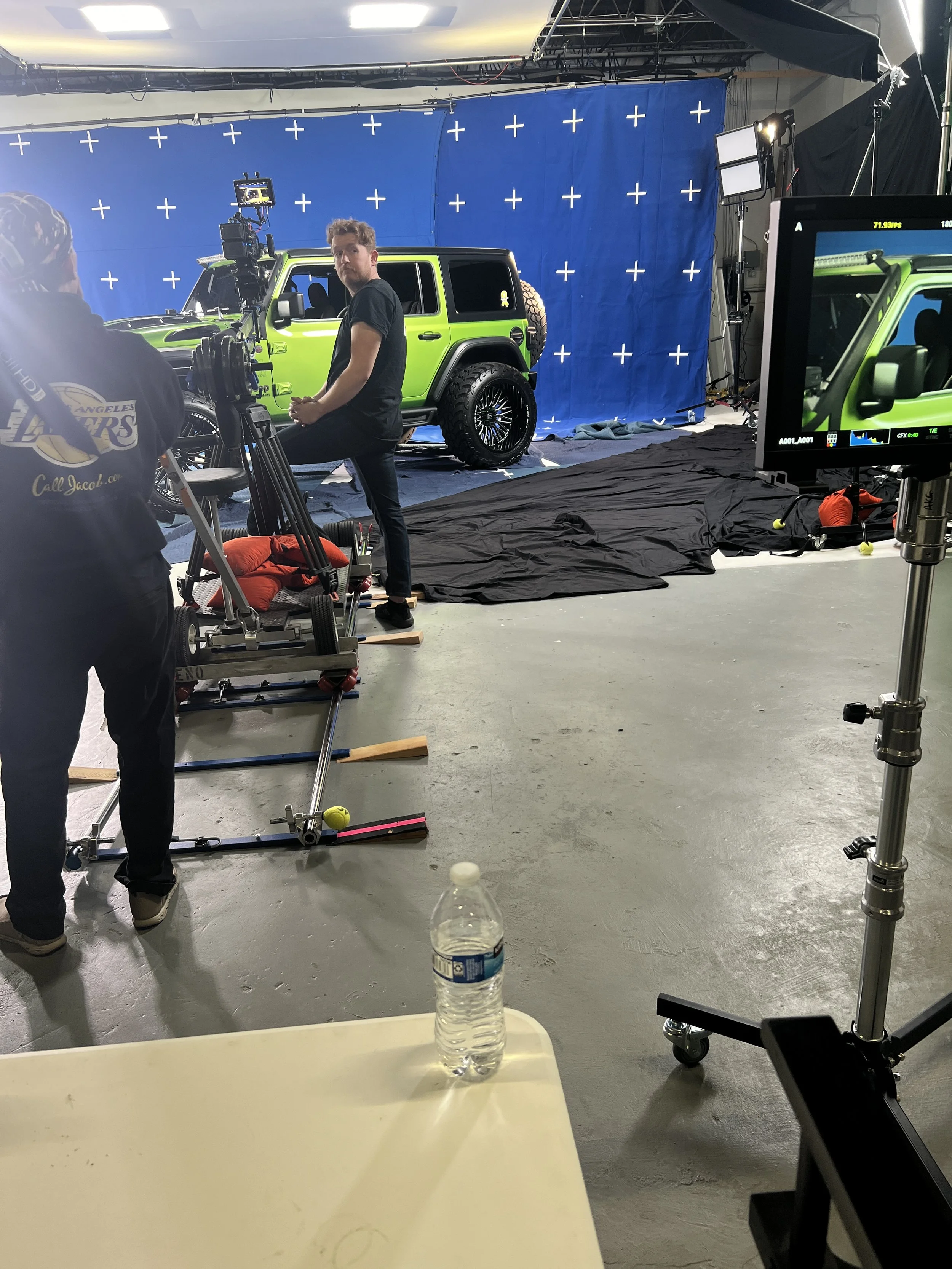 Inside a video production studio, a green Jeep with a blue screen backdrop is being filmed. Crew members and equipment are visible, including cameras, monitors, and lighting.