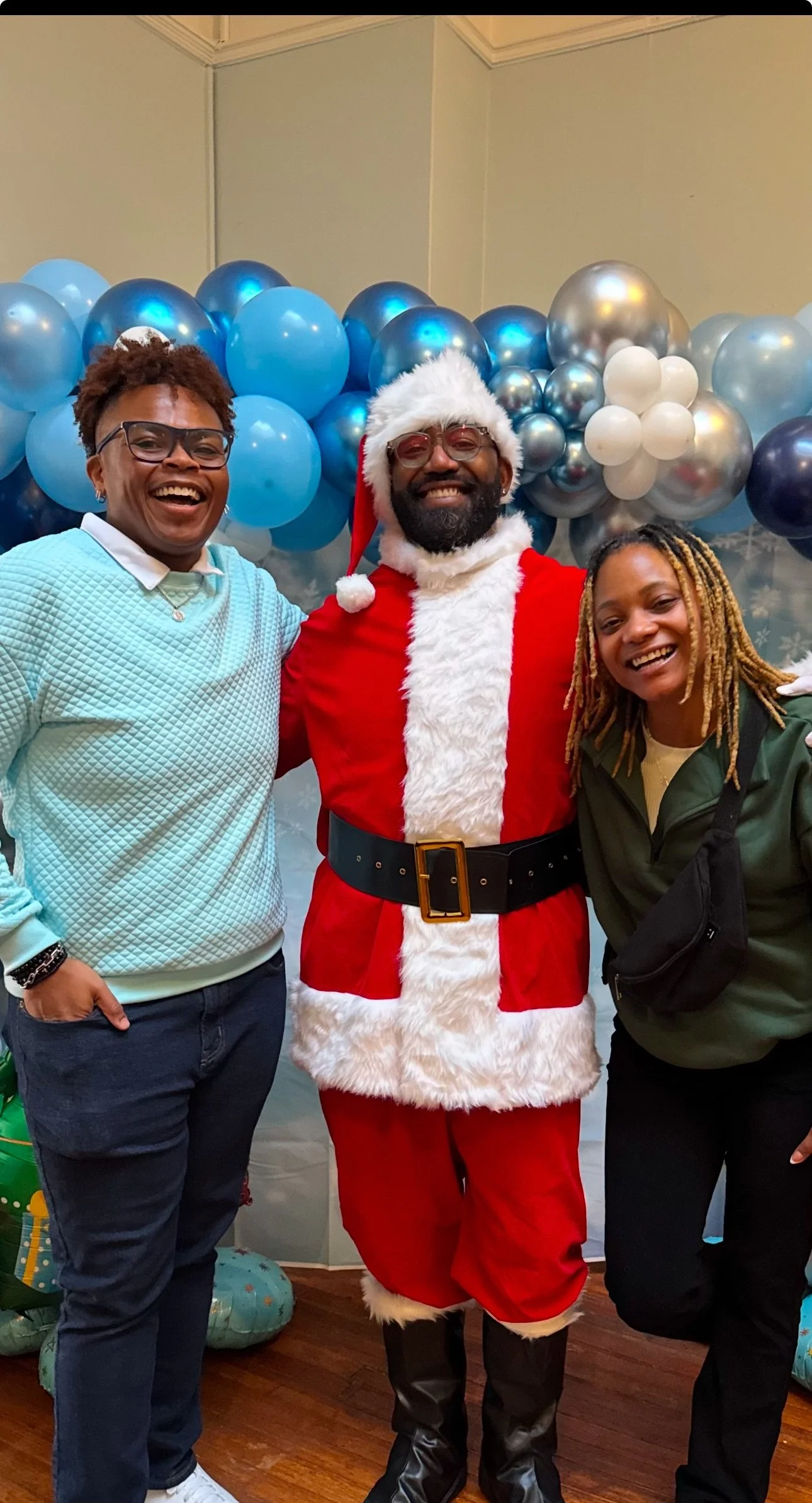 Two women and a man dressed as Santa Claus smiling in front of Christmas balloon decorations.