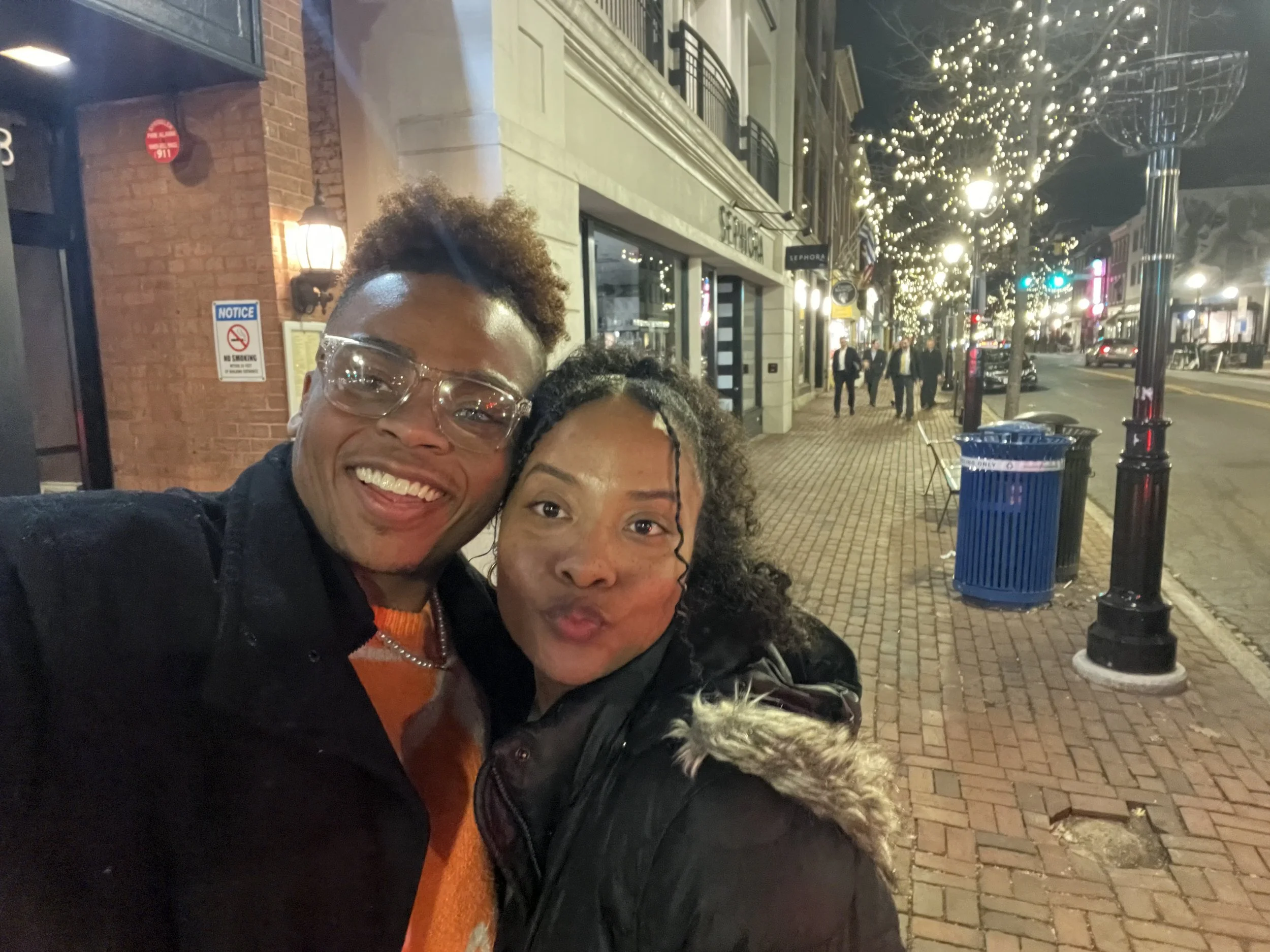 Two people taking a selfie on a city sidewalk decorated with string lights at night, with storefronts, people walking, and holiday lights in the background.