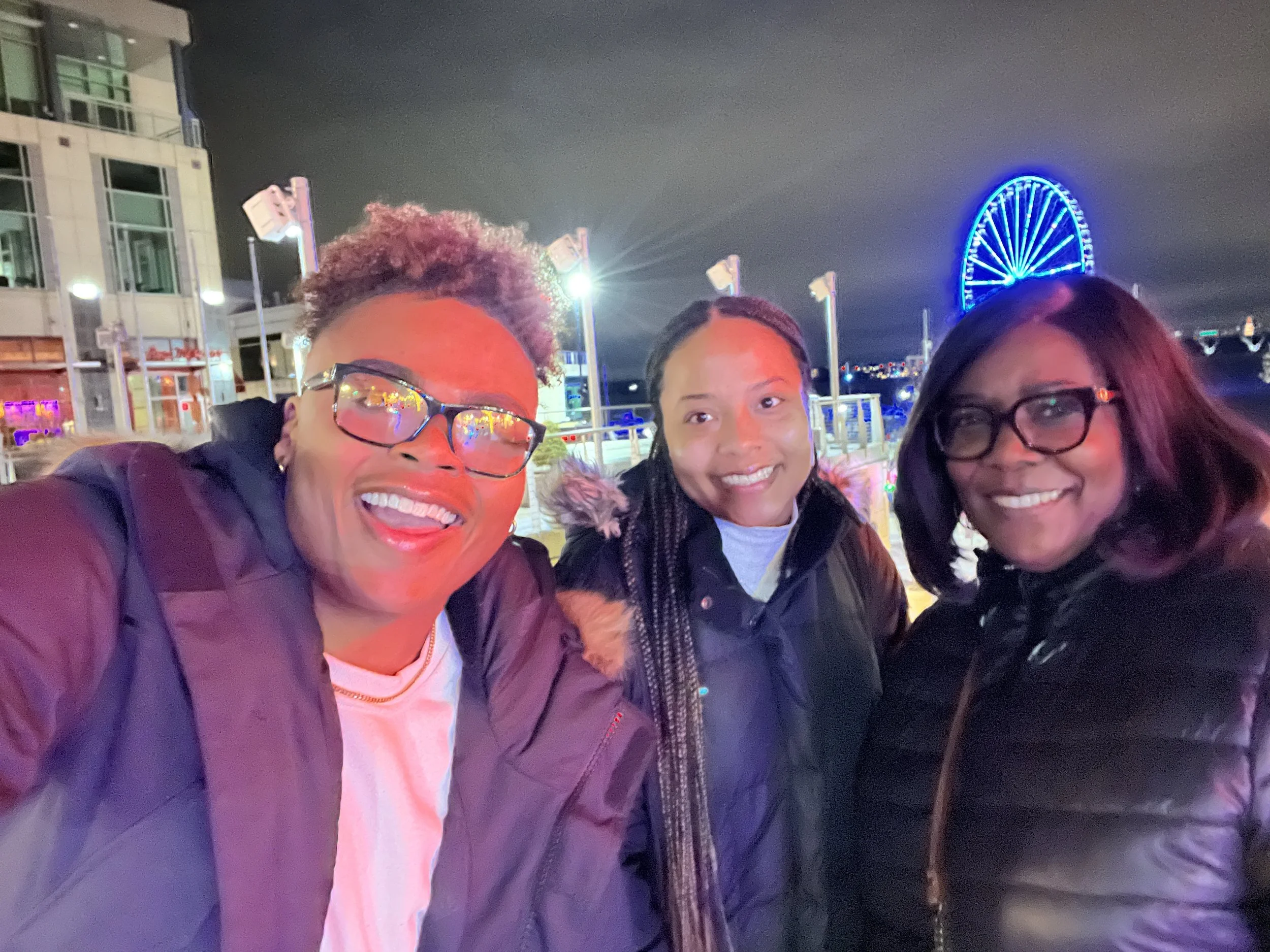 Three women smiling for a selfie at night near a city waterfront with a lit ferris wheel in the background.