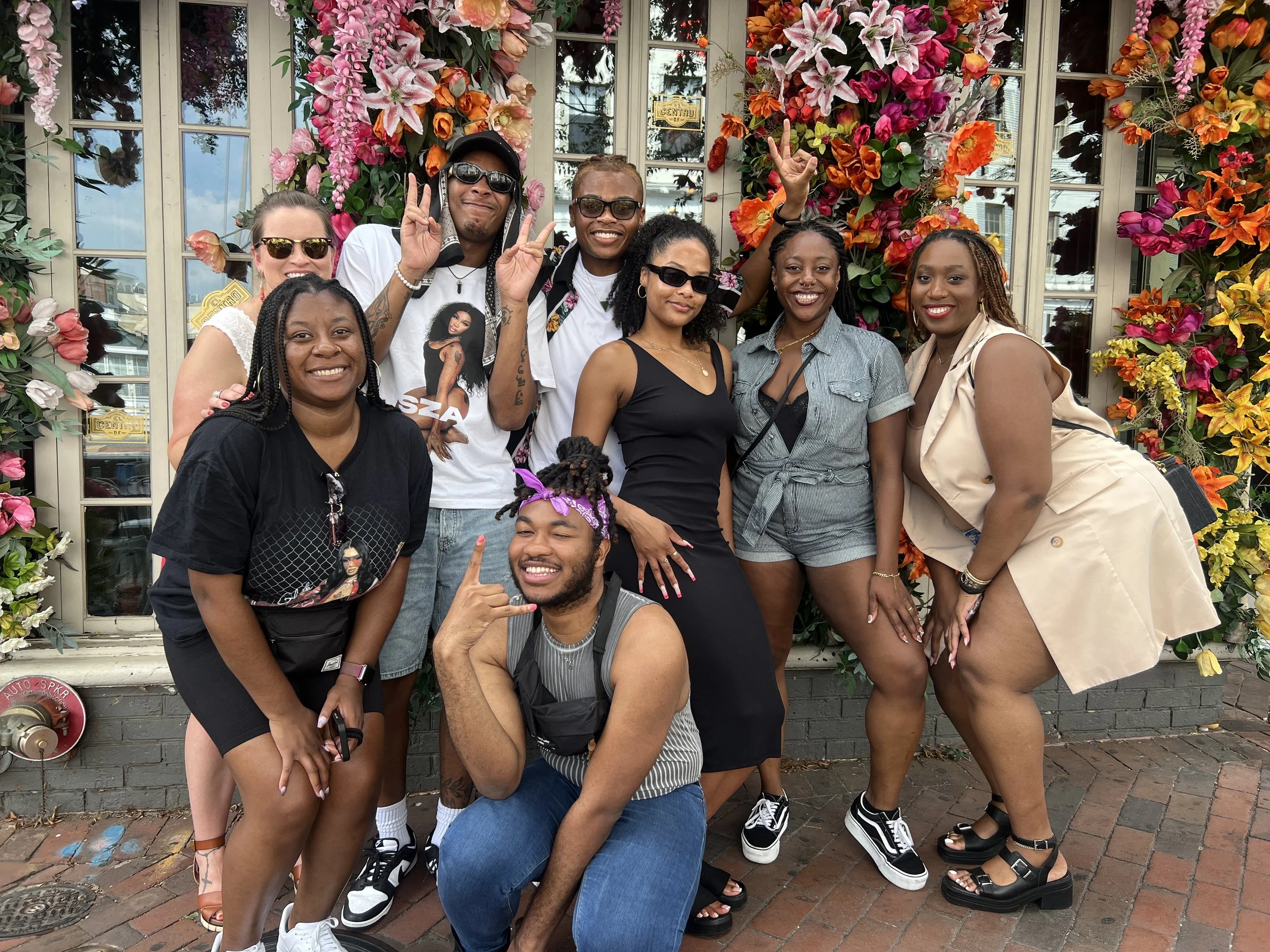 Group of nine diverse people posing in front of a colorful floral backdrop, smiling and making peace signs.