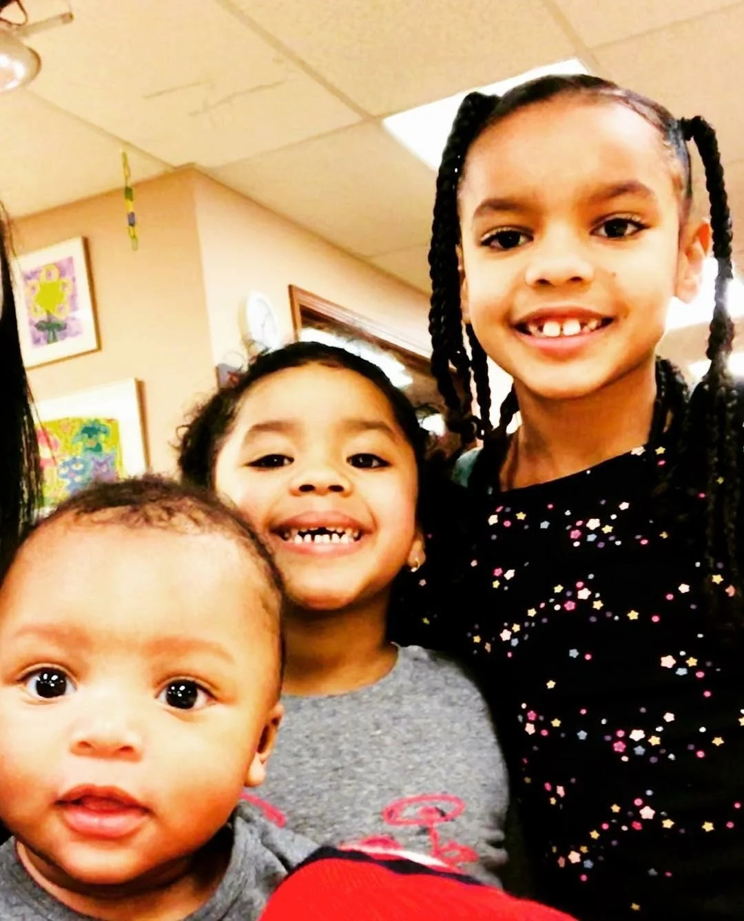 Three young children smiling in a community room with colorful artwork on the walls.