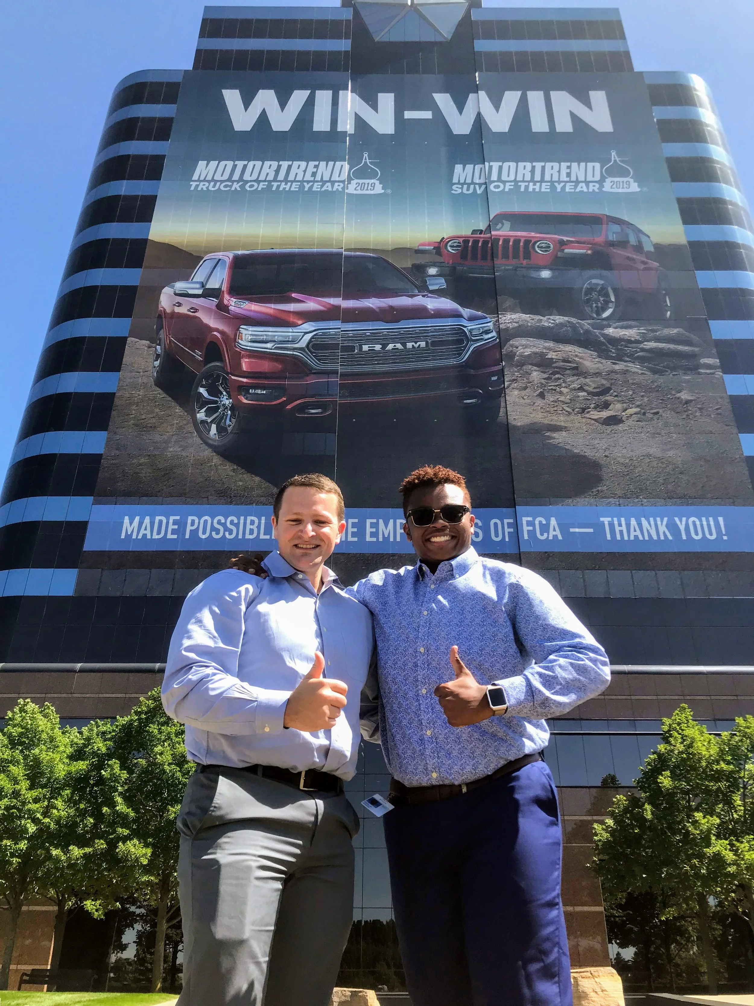 Two men smiling and giving thumbs up in front of a tall building with a large advertisement for RAM trucks, featuring images of a red truck and an SUV, with the text "WIN-WIN" and "MADE POSSIBLE BY THE EMPLOYEES OF FCA."
