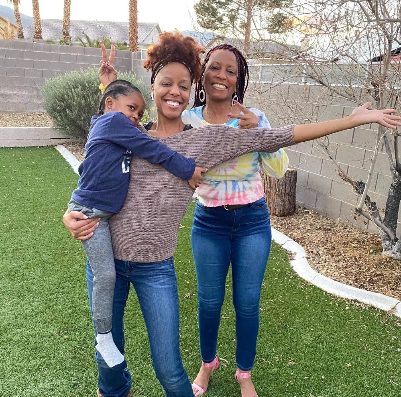 Three women and a young girl posing outdoors with a backyard background, smiling and making peace signs.