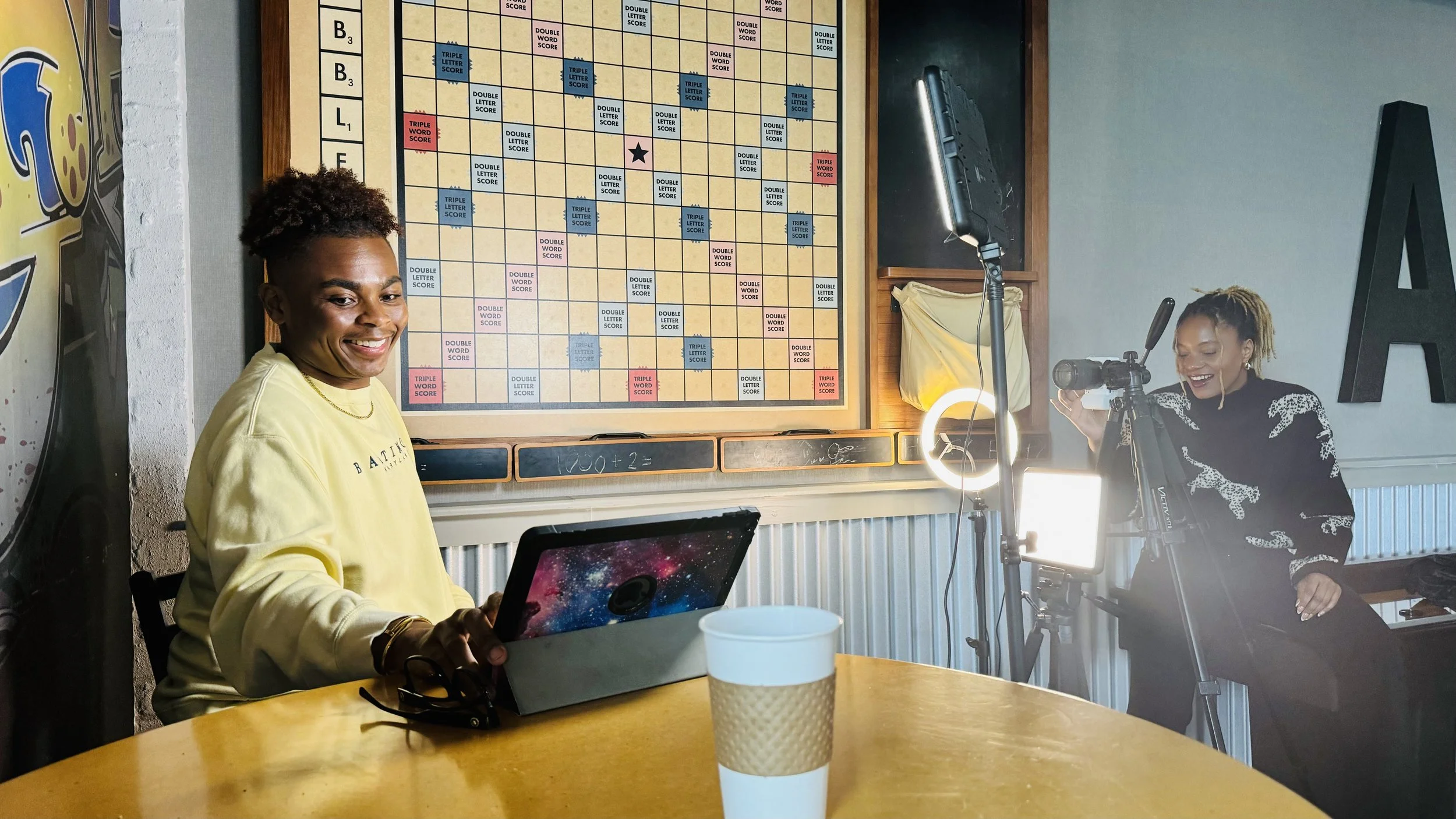 Ken sitting at coffee table, looking at tablet, infront of large scrabble board